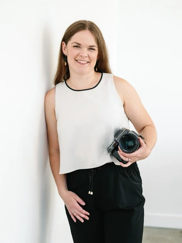 A woman smiling and holding a professional camera while standing against a white wall.