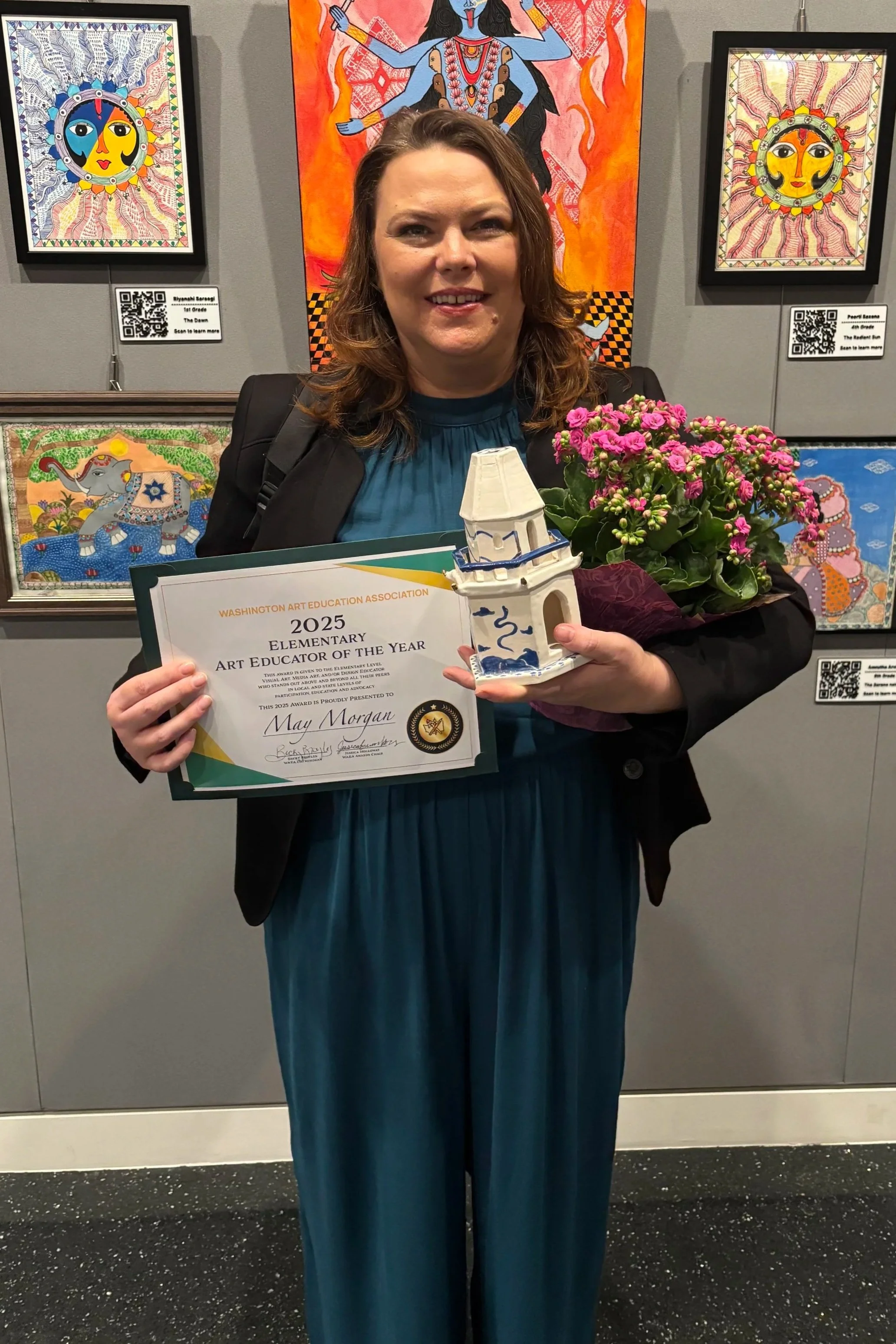 A smiling woman in a blue dress holding a small trophy and a certificate, standing in front of colorful artwork of animals and celestial themes, next to a pink flowering plant.