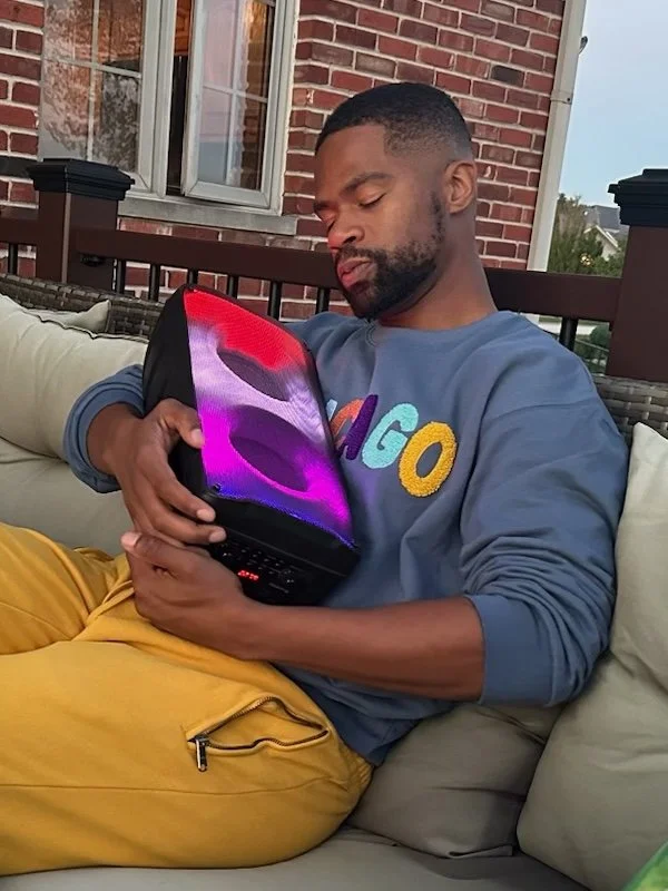 A man relaxing on an outdoor couch, wearing a blue hoodie with colorful letters and mustard yellow pants, holding a laptop with a vibrant screen.