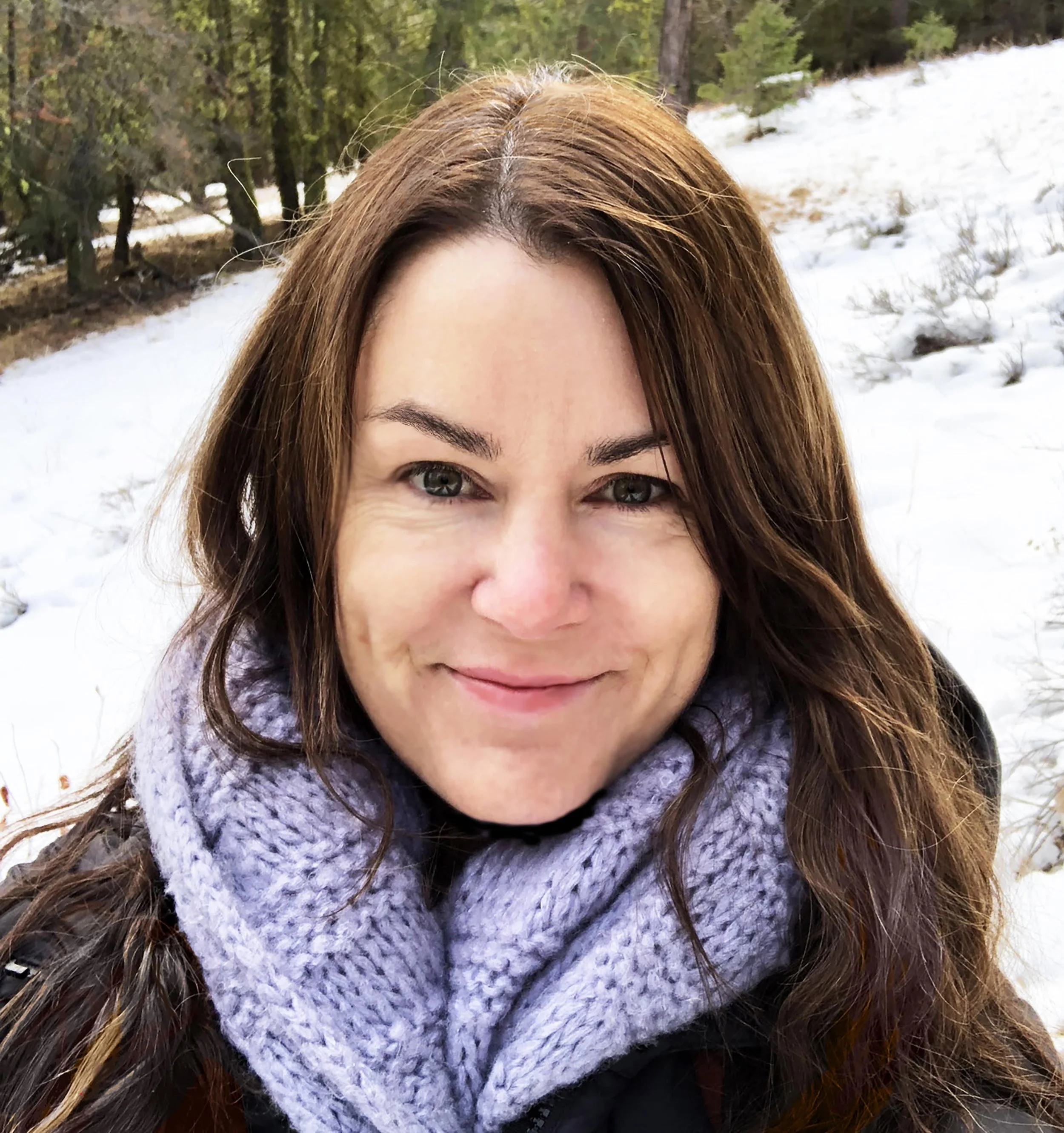Close-up of a woman with long brown hair and green eyes, smiling outdoors during winter, wearing a purple knit scarf, with a snowy landscape and evergreen trees in the background.