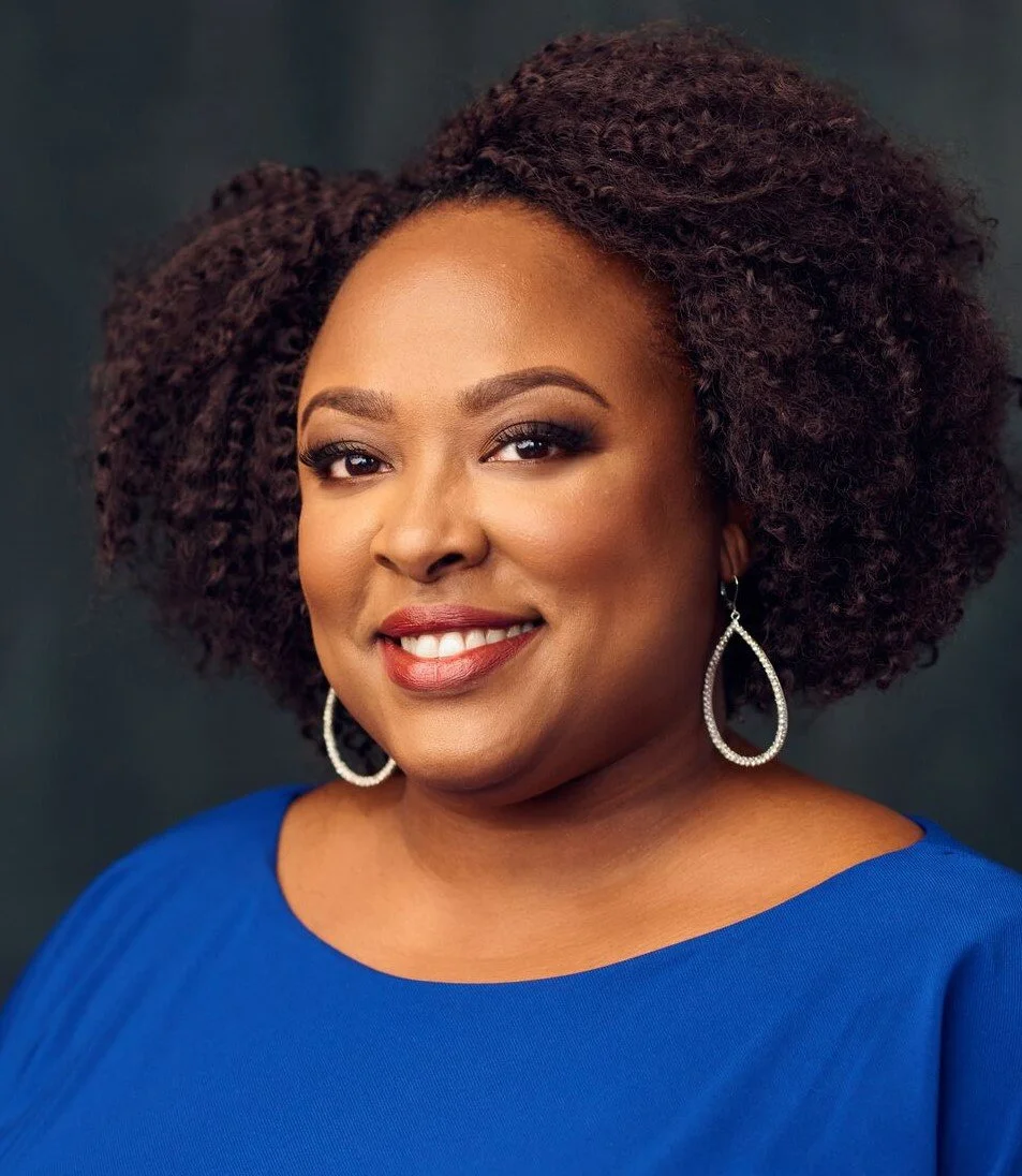 A woman with a curly hairstyle smiling, wearing a blue top and large teardrop-shaped earrings.