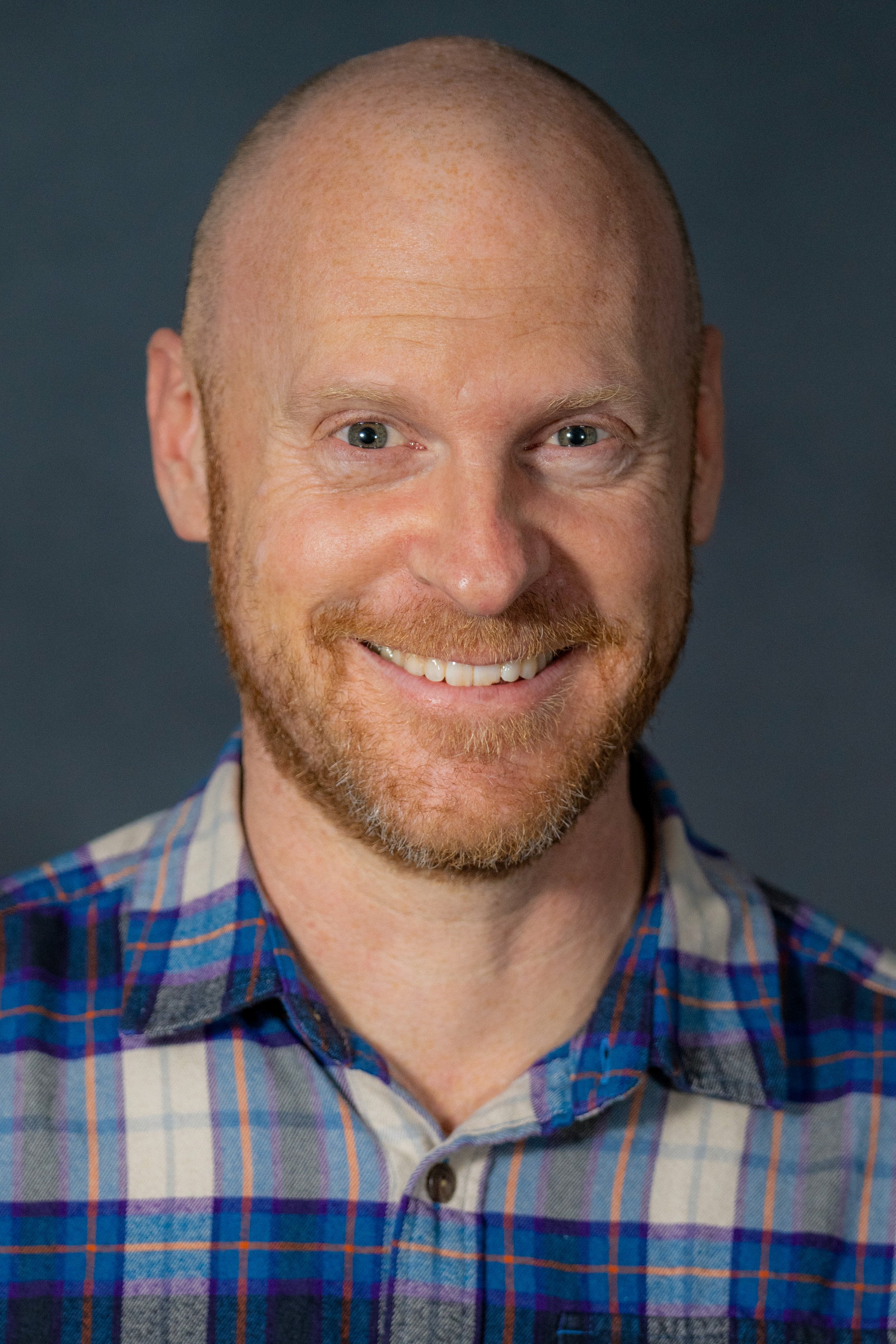 Close-up portrait of a smiling man with a beard and short hair, wearing a blue and plaid button-up shirt, against a dark background.