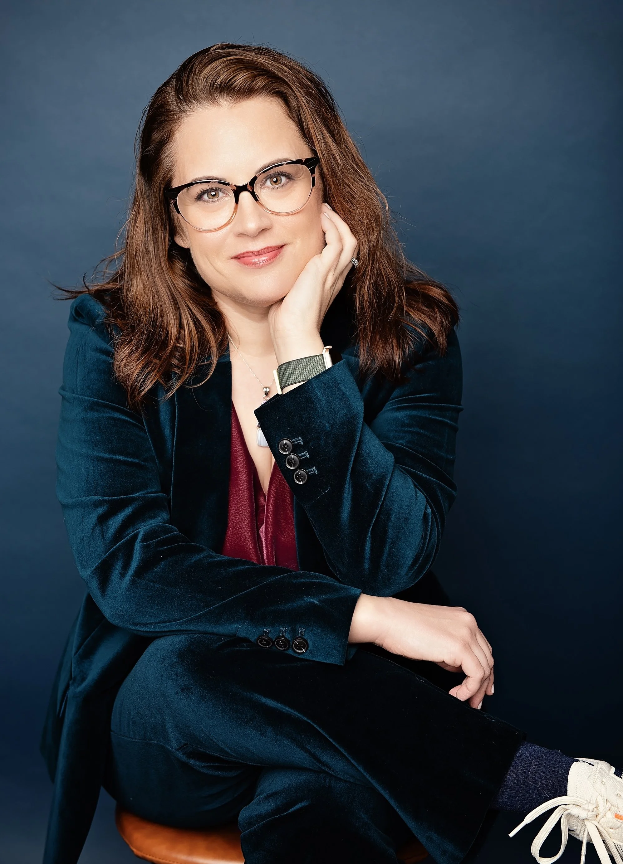 A woman with red hair wearing glasses sitting on a wooden stool with a dark blue background. She is dressed in a dark blue velvet blazer, light-colored shirt, black pants, and white sneakers. She has a gentle smile, resting her chin on her right hand, and looks directly at the camera.