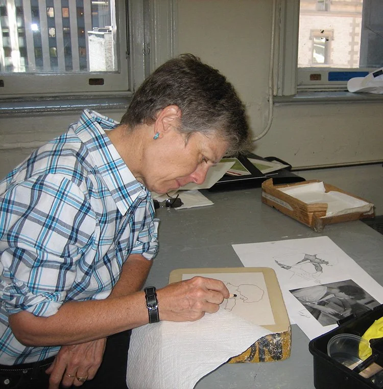 Artist Brenda Berkman sketching a drawing on a lithograph stone with papers and art supplies.