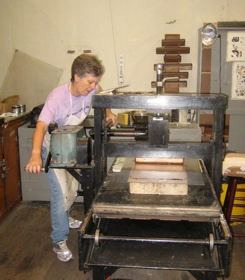 The artist Brenda Berkman printing her artwork standing next to a printing press in a workshop, with tools and equipment nearby.