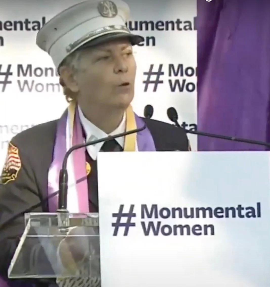 Brenda Berkman in her FDNY dress uniform with women's suffrage scarf, speaking at a podium with a sign that reads 'Monumental Women' at the unveiling of the Central Park Women's Rights Pioneers Monument 2020.