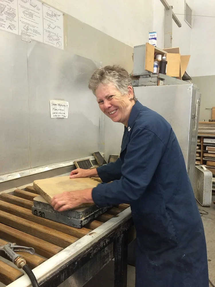 Artist Brenda Berkman in her blue work coat is smiling while grinding a lithograph stone in the print shop in preparation for creating a new lithograph print.