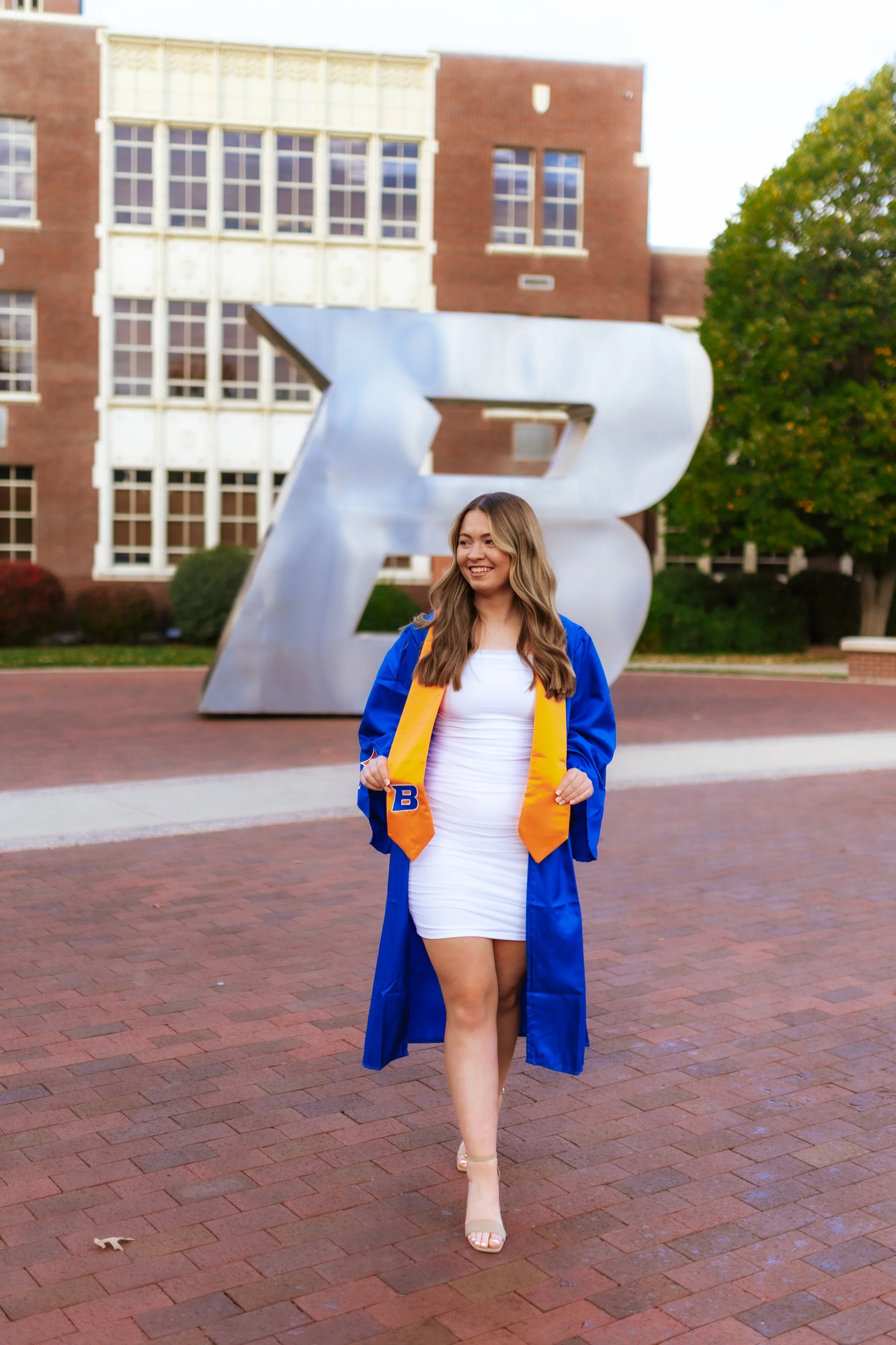 A young woman in a white dress and blue graduation gown with a yellow stole walking on a brick pathway, smiling. Behind her is a large metallic letter 'B' sculpture and a brick building with large windows, trees, and clear skies.