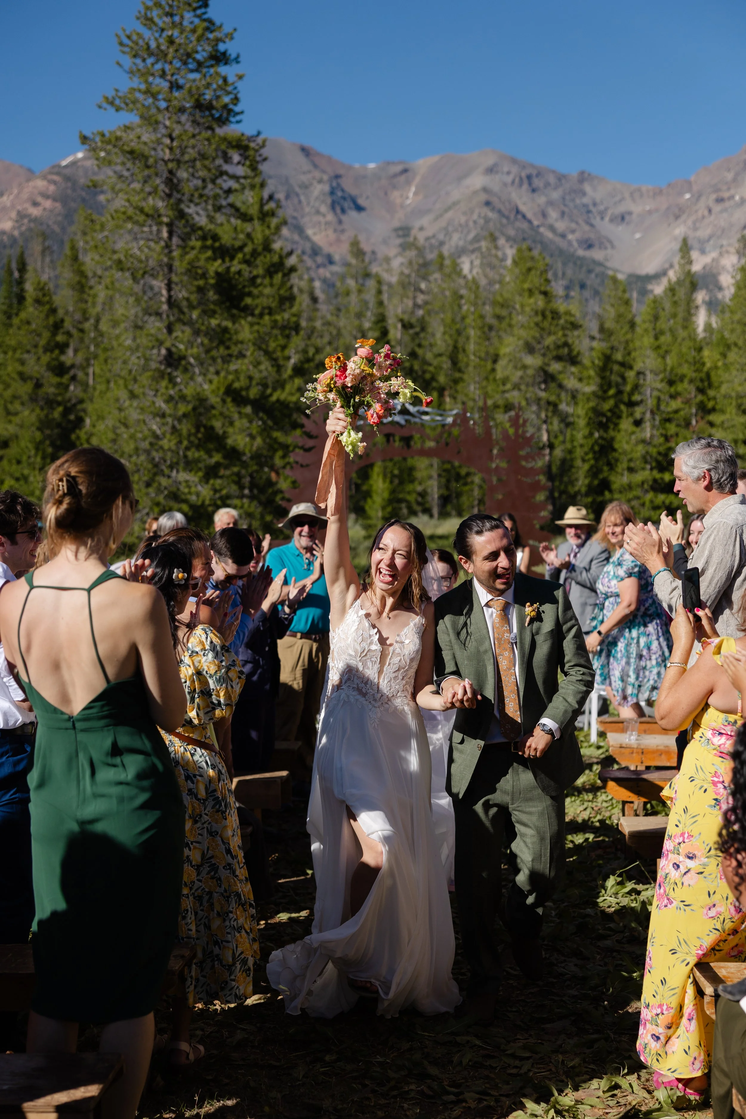 A newlywed couple walking down the aisle outdoors, surrounded by smiling guests, with the bride holding a bouquet of flowers high in the air, against a backdrop of mountains and trees on a sunny day.