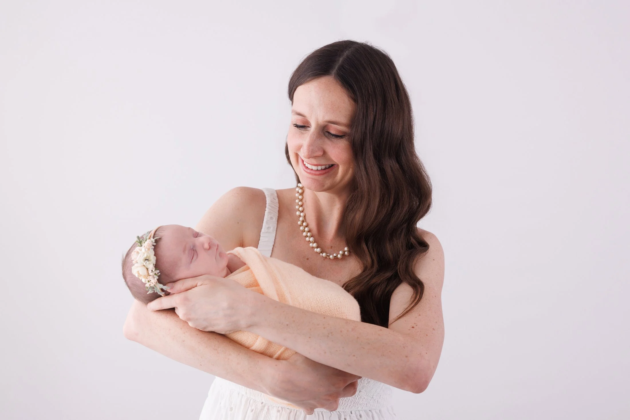 mom in a white dress with pearls holding newborn baby and looking and smiling at her