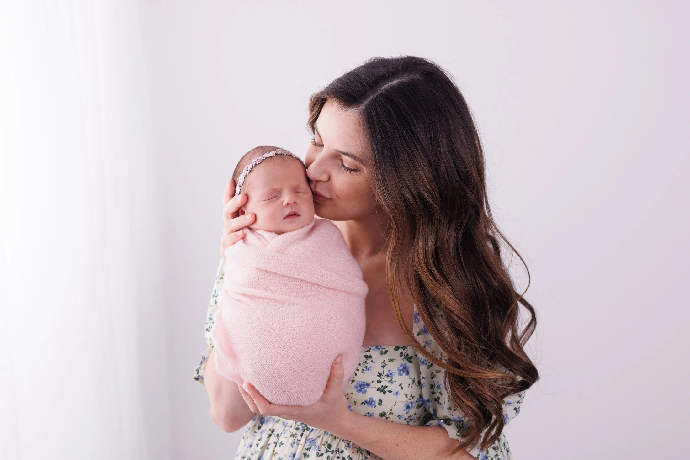 mom in a floral dress kissing newborn baby in a pink wrap
