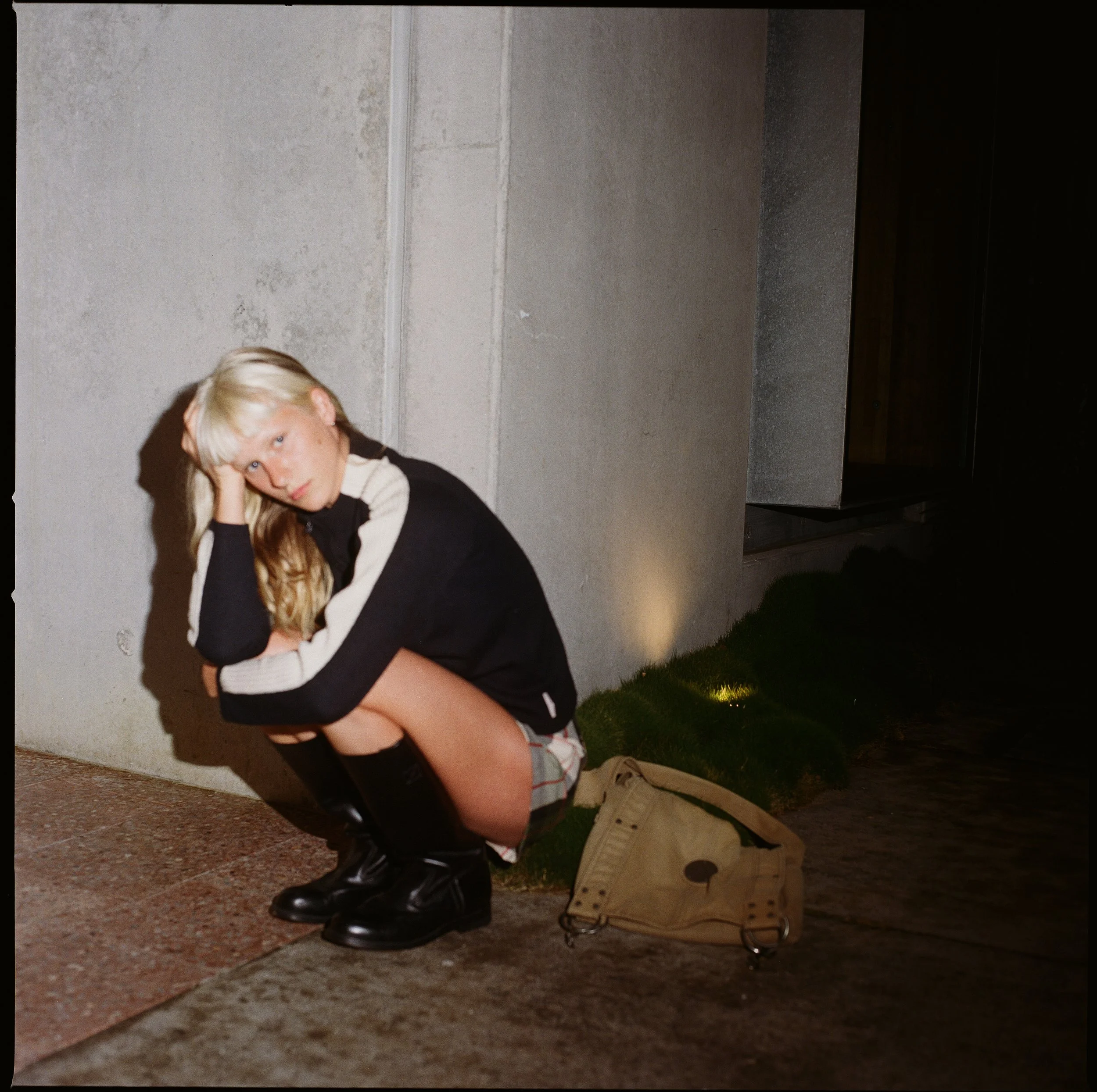 A young person with blonde hair and light skin sitting on the ground, resting their head on one hand, wearing a black and white sweater, black shoes, and tan pants. There is a beige bag on wheels nearby, with a gray ribbed wall behind them and some grass in the foreground at night.