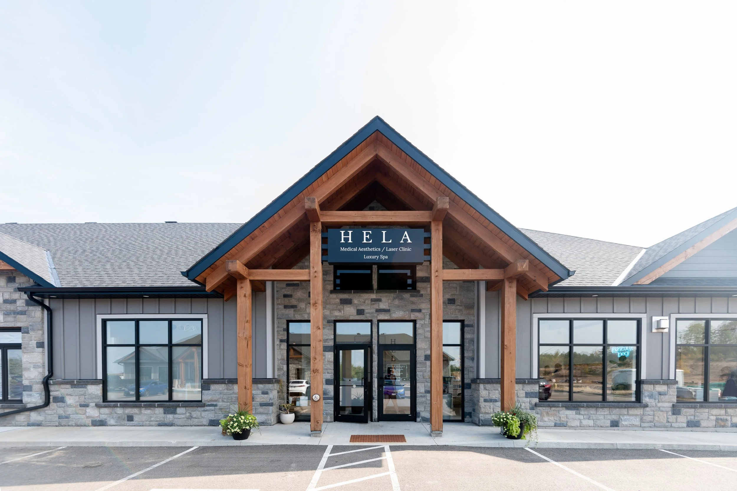 Front view of a modern medical spa and laser clinic named HELA with a stone and wood exterior, large windows, and a parking lot in front.
