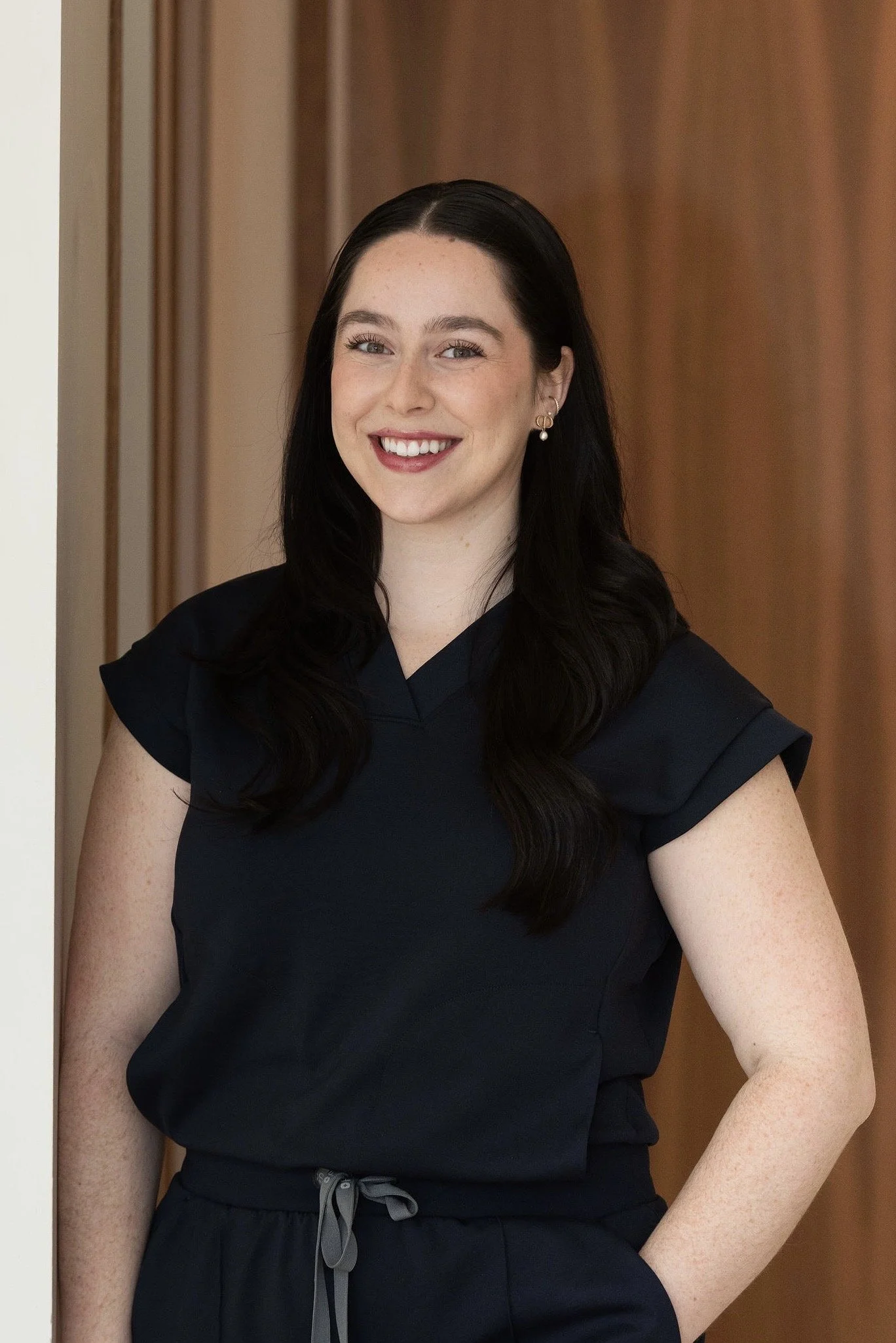 A young woman with long dark hair, wearing a taupe medical scrub top, smiling and leaning against a wall in a professional setting.