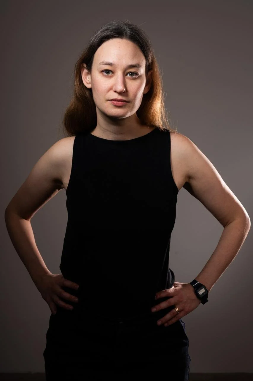 A person with short dark hair and light skin is outdoors in a wooded area, looking to the right. They are wearing a black t-shirt and carrying a backpack. Bright sunlight filters through the green leaves of the trees.