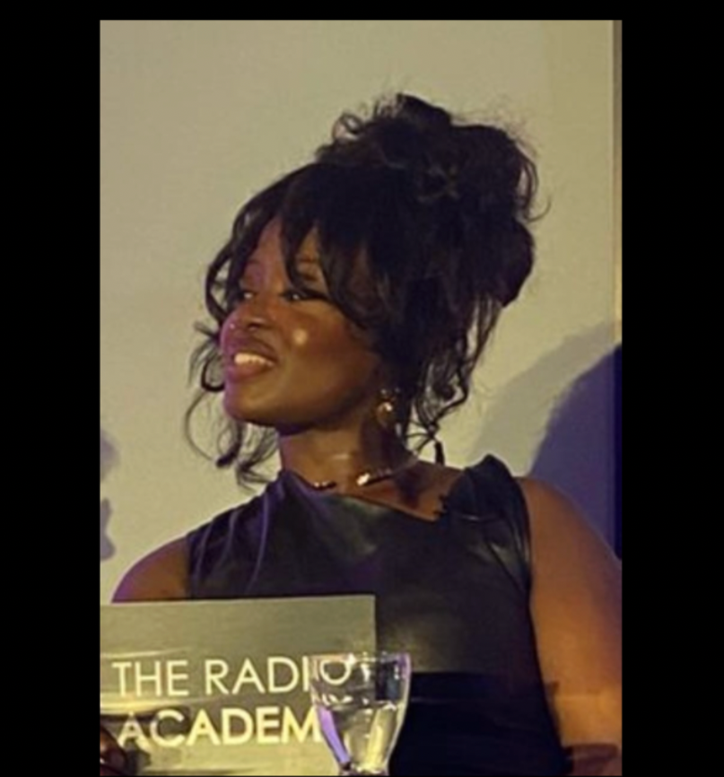 A woman with braided hair and earrings, looking slightly to her left, in a room with a plant and framed artwork in the background.