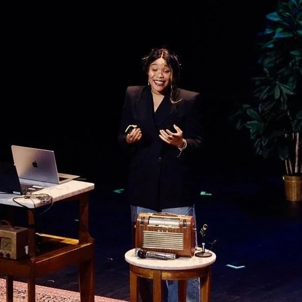 Portrait of a woman with shoulder-length black hair wearing a white button-up shirt, standing outdoors with a blurred background of a boat and sky.