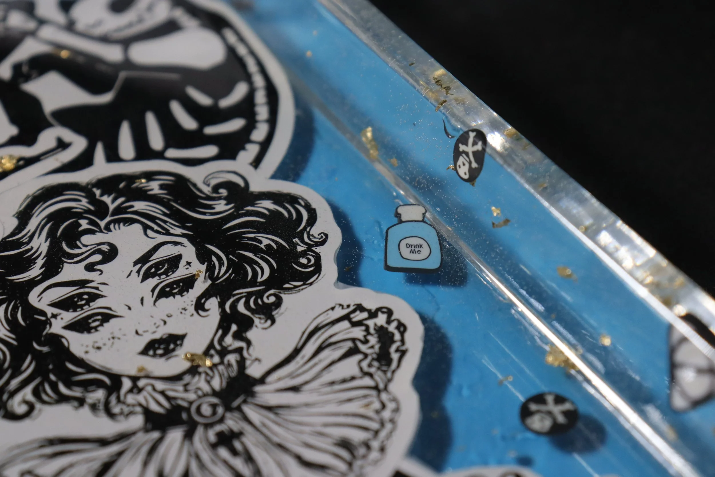 Close-up of a sticker with a black and white intricate illustration of a woman's face and hair. Part of the sticker shows a small cartoon-like bottle labeled 'Drink Me' with a sponge bob style face and pirates hat. The sticker is placed on a clear su