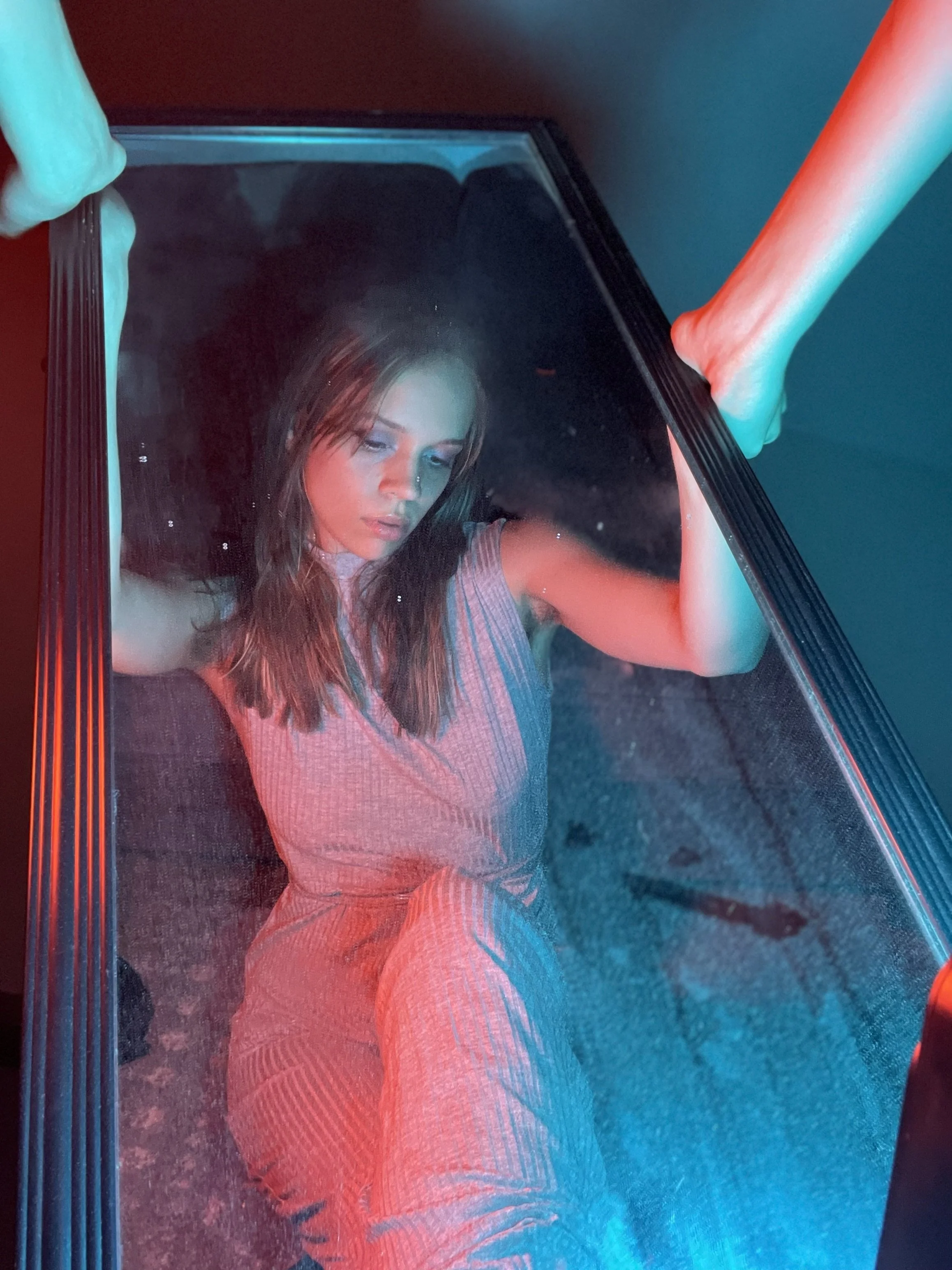 Young woman with long hair wearing a striped shirt, taking a photo with a smartphone, reflected in a mirror with neon lighting.