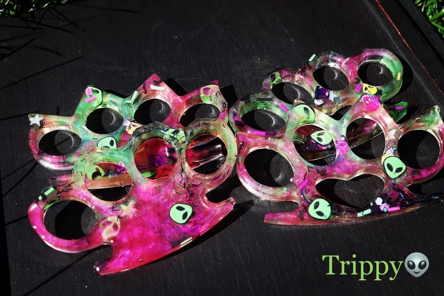 Colorful pair of brass knuckles with green, pink, and purple swirls and alien face stickers on a black surface.
