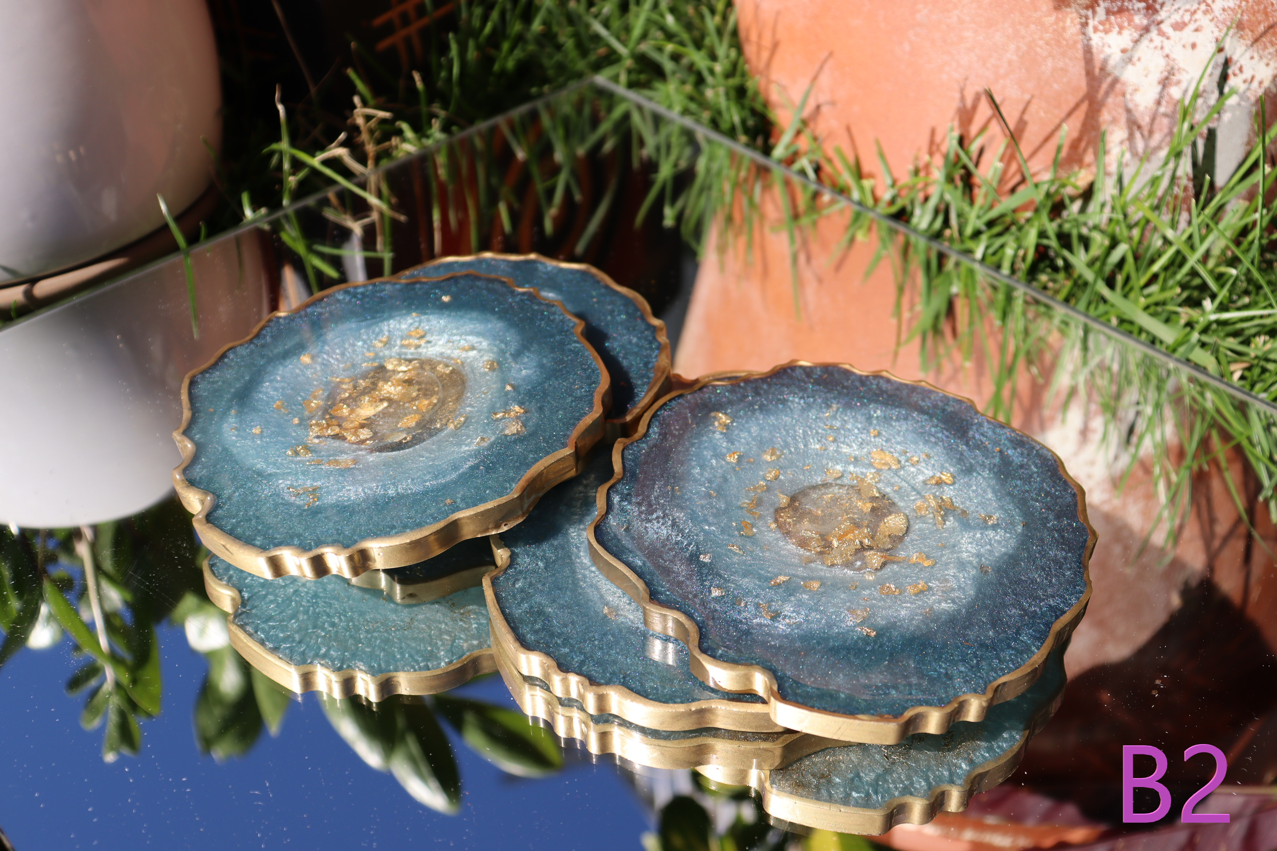 Close-up of three tiered resin and metal coasters with gold flakes, outside on a reflective surface, with green grass and brick in the background.