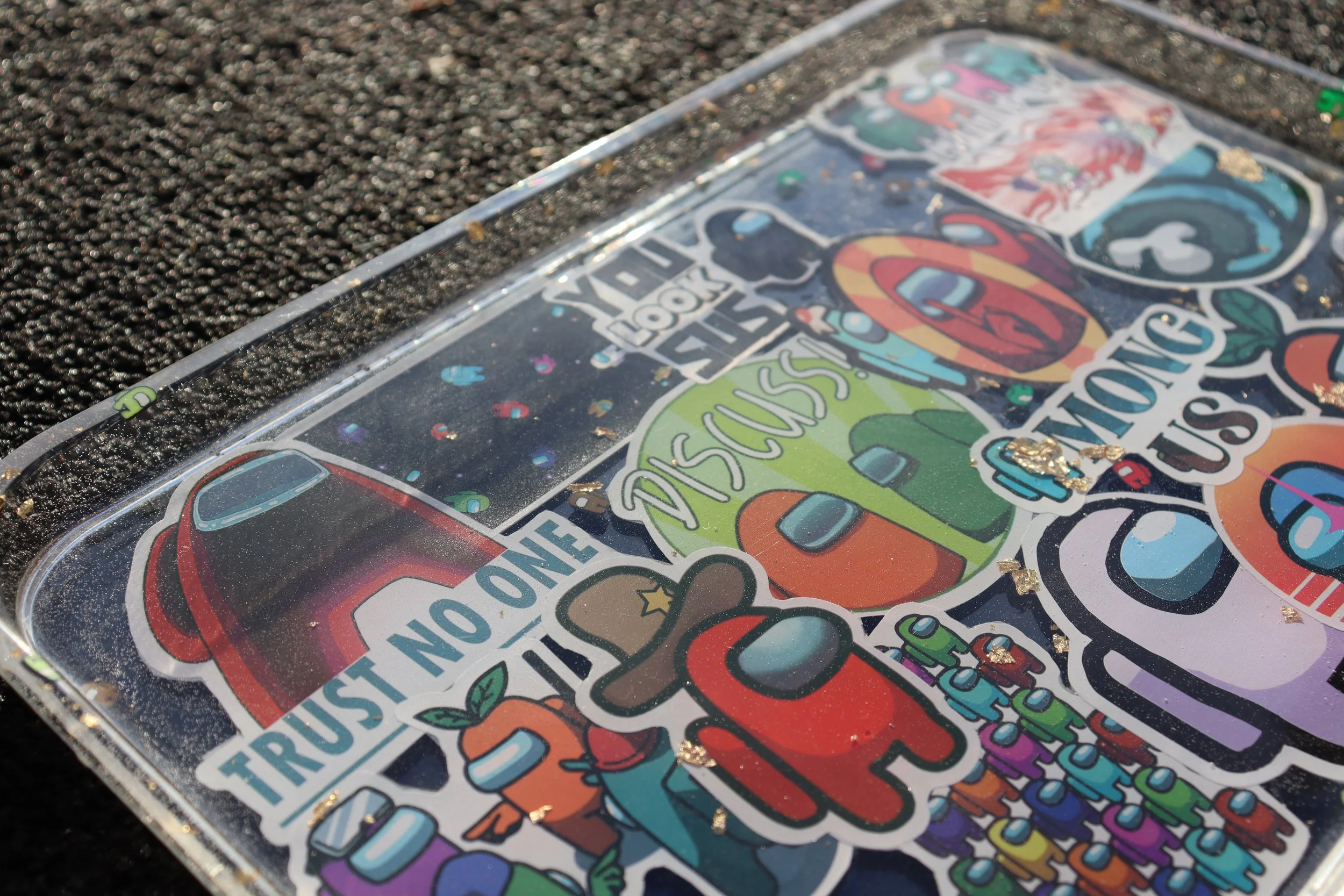 Close-up of a plastic tray with colorful stickers featuring the Among Us characters and phrases like "Trust" and "Discover", placed on asphalt.