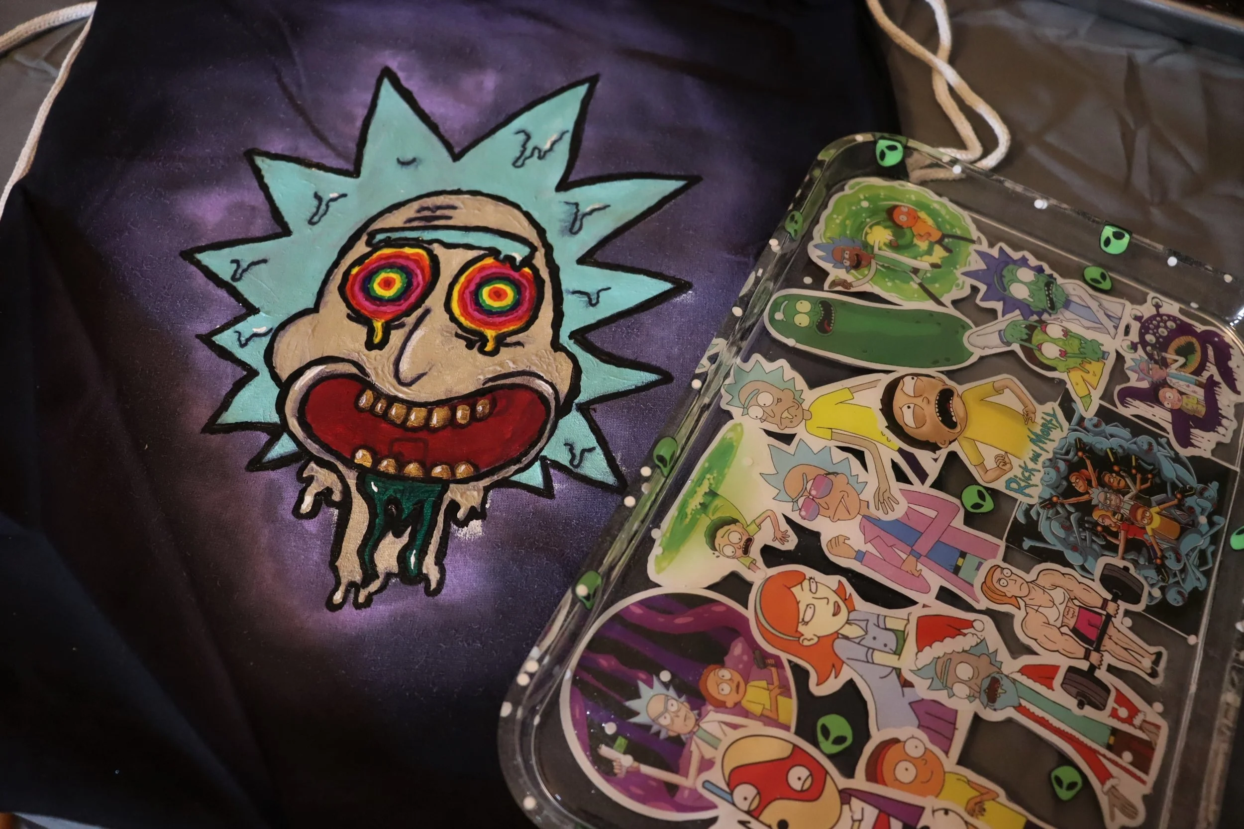 A black drawstring bag with a colorful graphic of Rick Sanchez from Rick and Morty, featuring a distorted face with spiraled eyes and a wide red mouth. Next to the bag is a collection of Rick and Morty-themed stickers, some with alien faces and vario