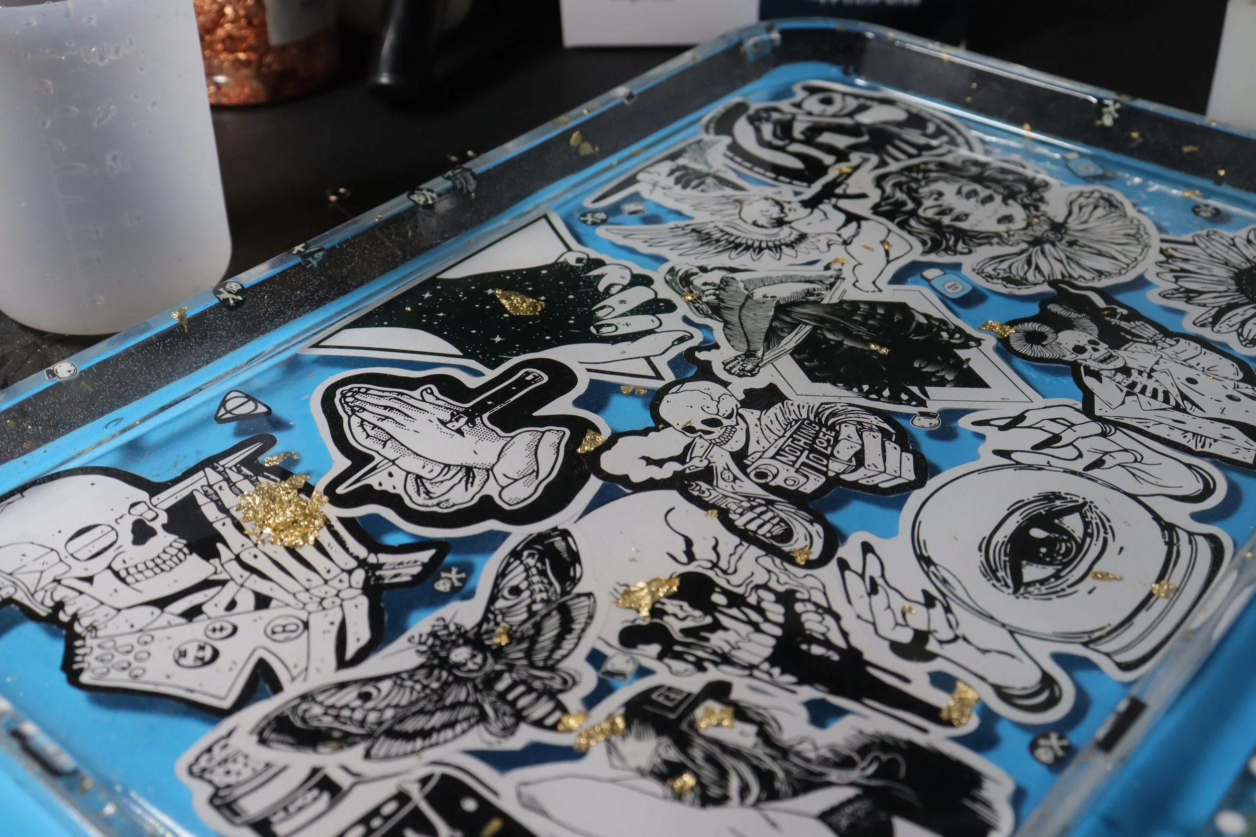 Close-up of a surface covered with various black and white tattoo-style stickers featuring skulls, hands, animals, and abstract designs, with some gold glitter scattered on top.