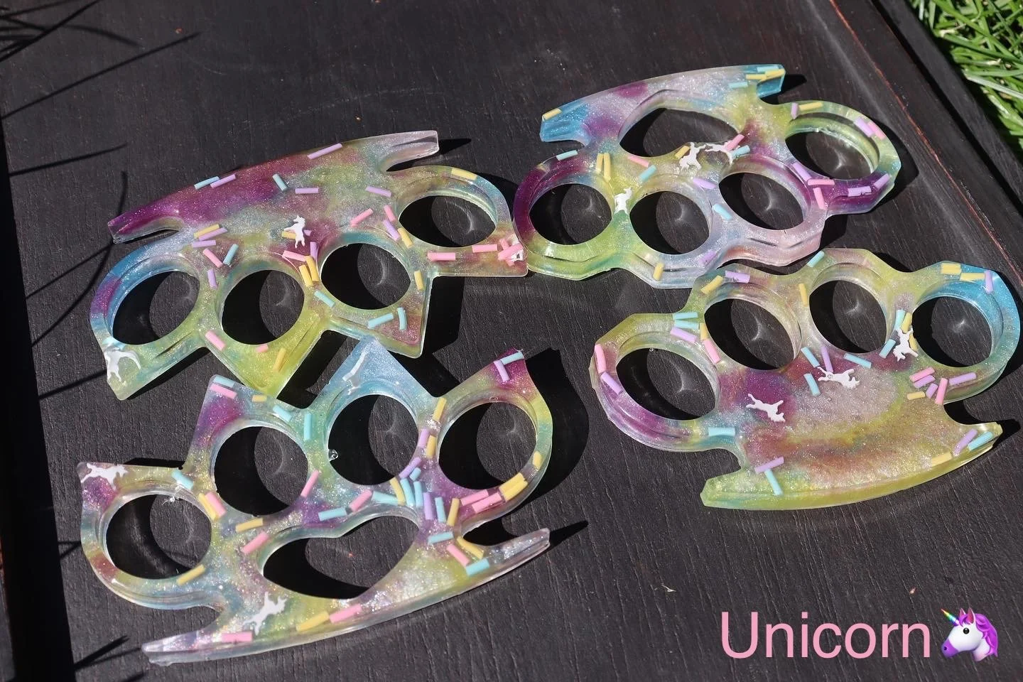 Colorful rainbow unicorn-shaped sunglasses with sprinkles, lying on a dark surface, labeled 'Unicorn' with a unicorn head emoji.