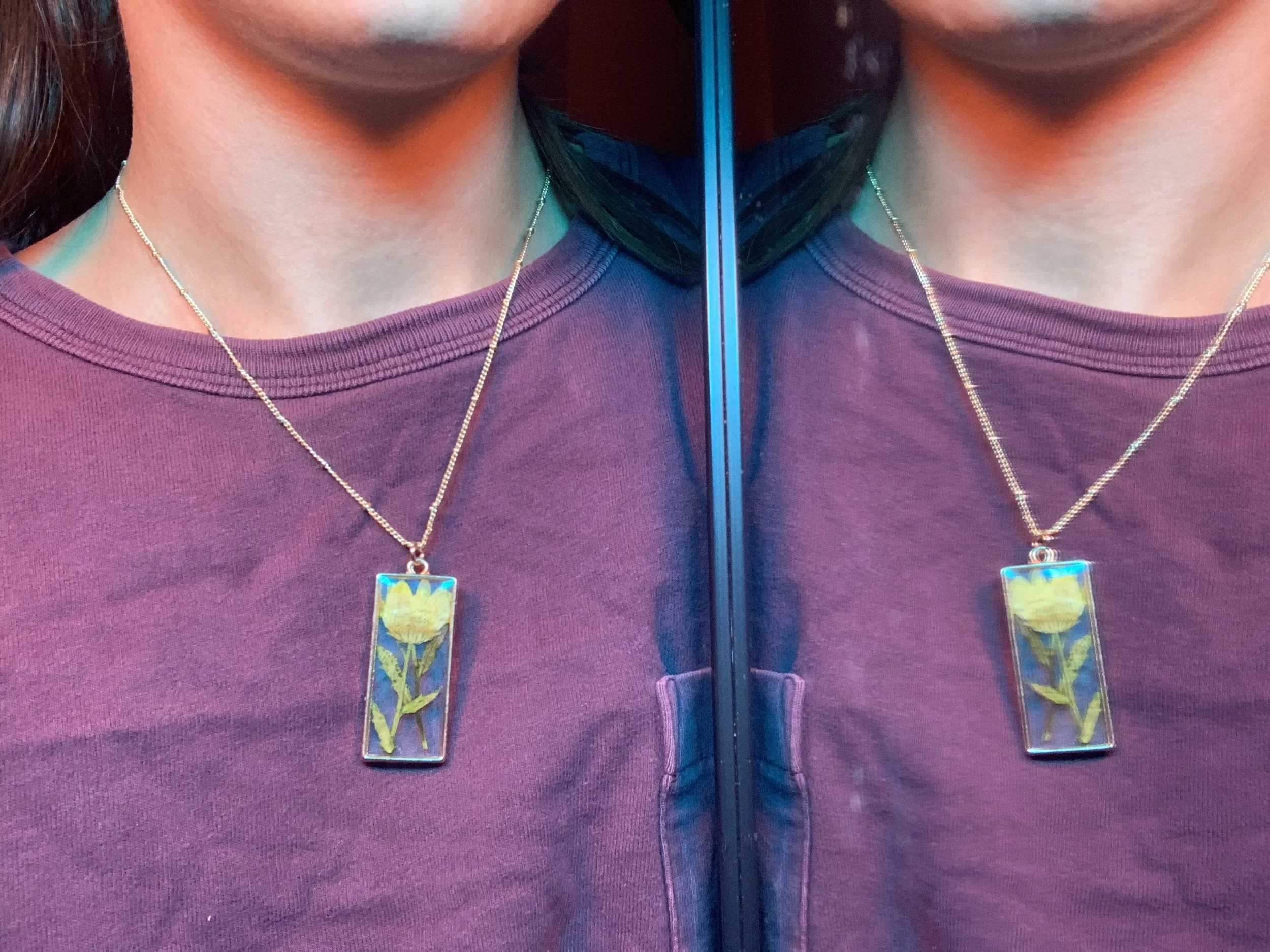 Two women are wearing purple shirts and necklaces with glass pendant charms showing yellow flowers. The image is split in half with a mirror reflection effect.