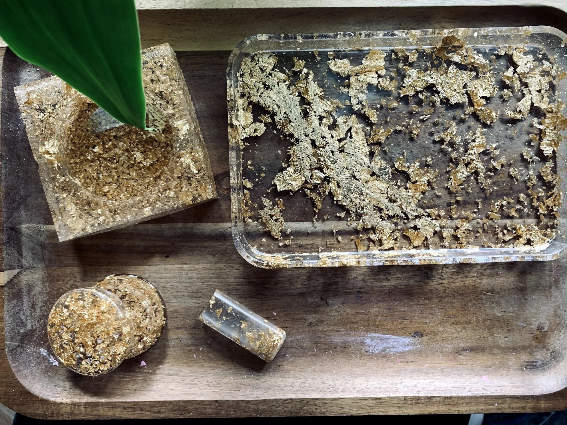 Gold leaf and resin craft materials on a wooden surface, with a small glass container and a green leaf.