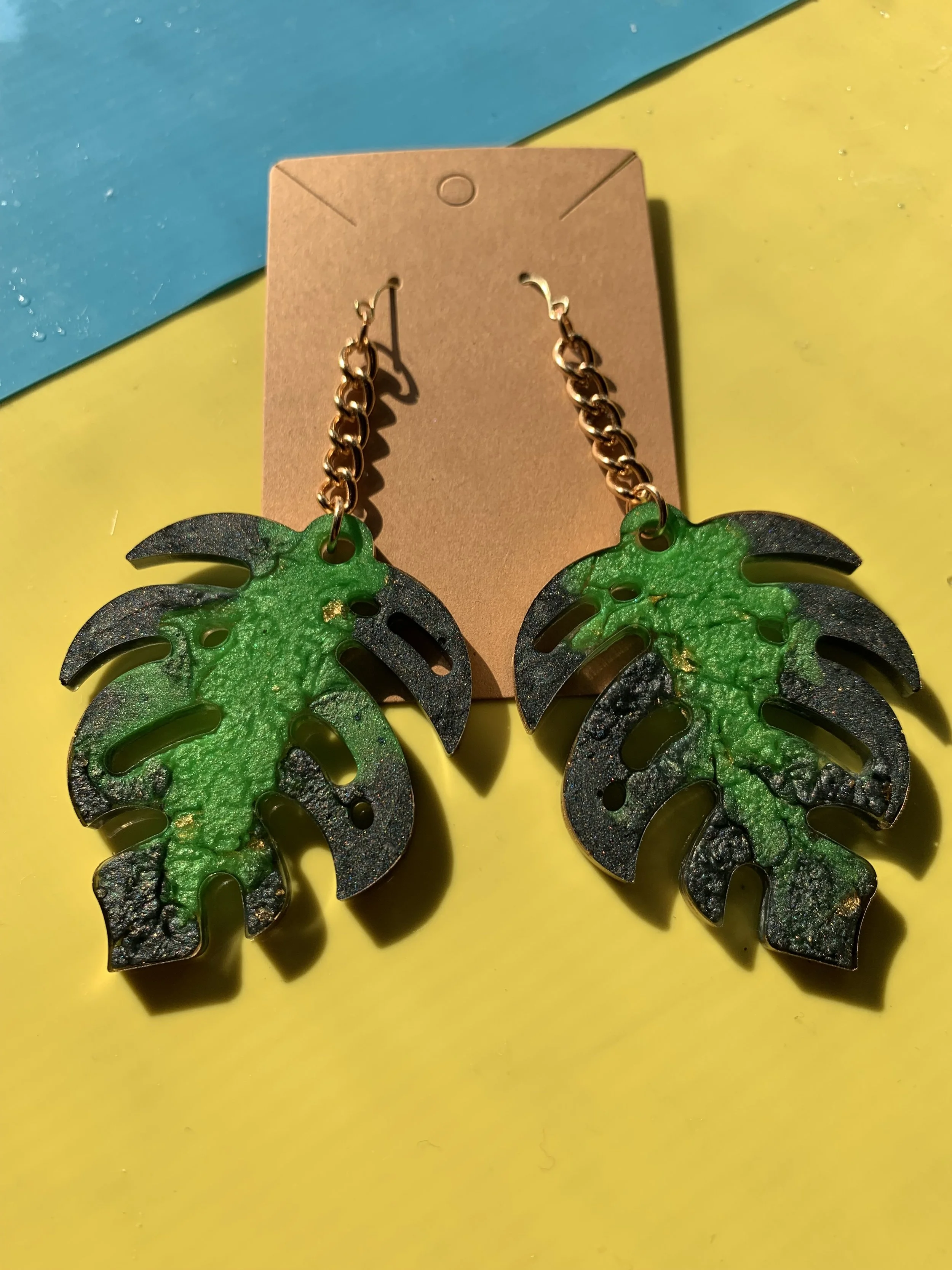 Green and black monstera leaf shaped earrings with gold chain hooks against a yellow and blue background.