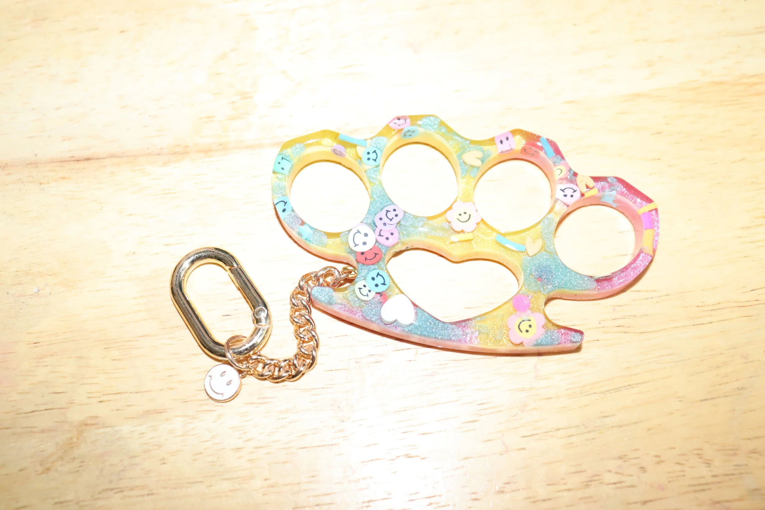 Colorful scratch-off keychain shaped like a bear paw with small smiley face emojis, accompanied by a metal keyring and a chain, on a light wooden surface.