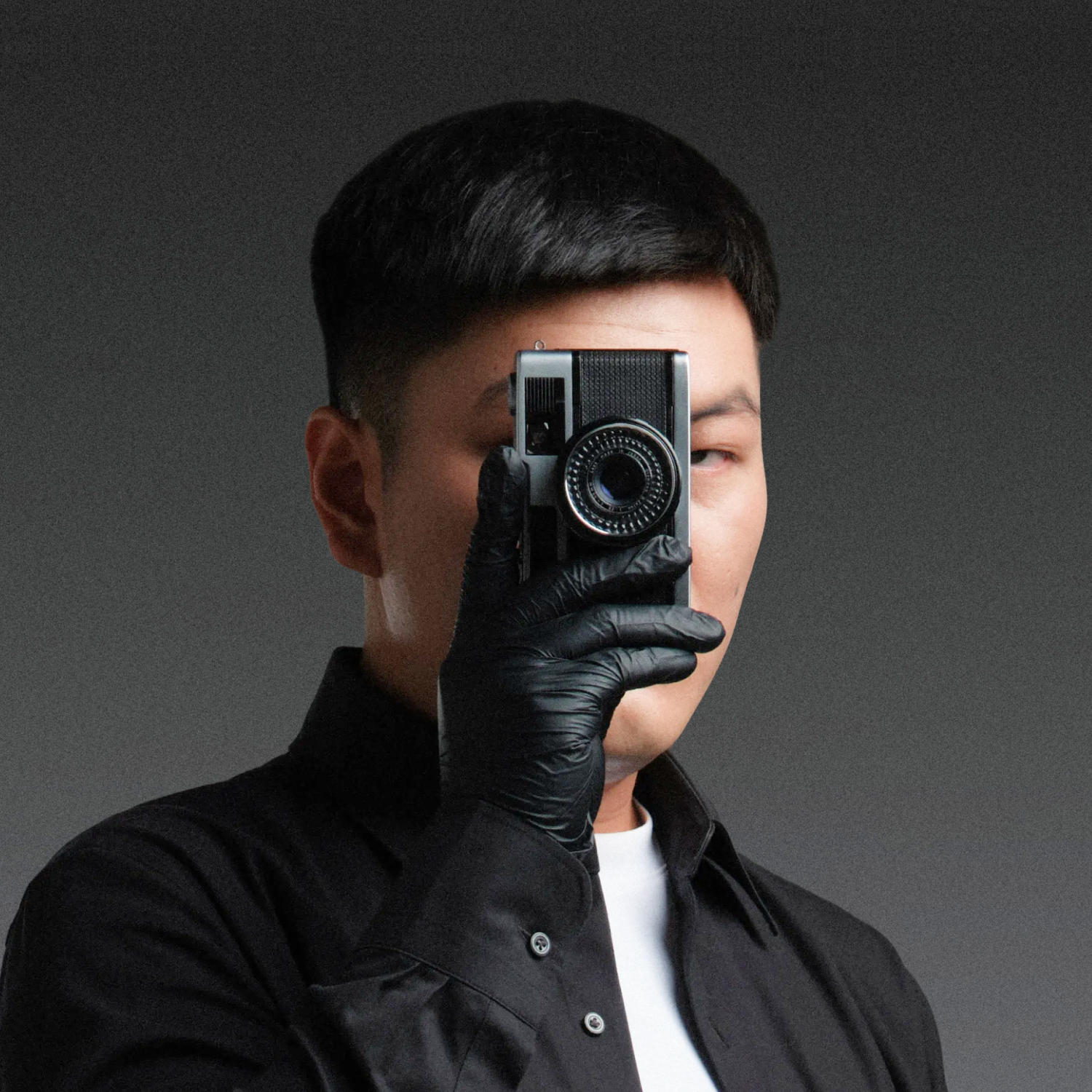 Person holding a camera, taking a photo, wearing a black shirt, with a plain gray background.