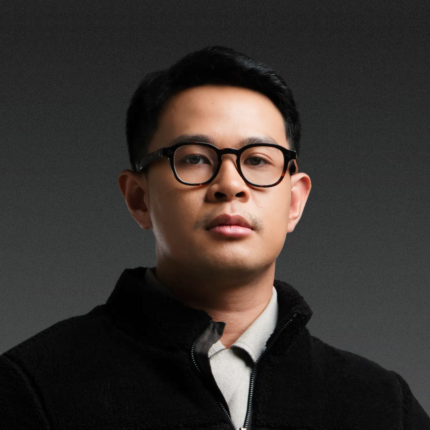 Man with black glasses and plaid shirt smiling at the camera, against a neutral background.