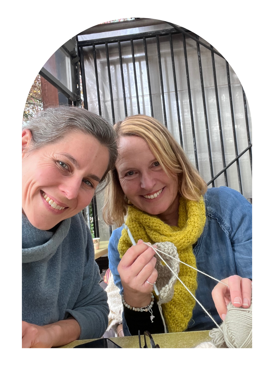 2 women smiling into camera, one holding yarn and knitting needles