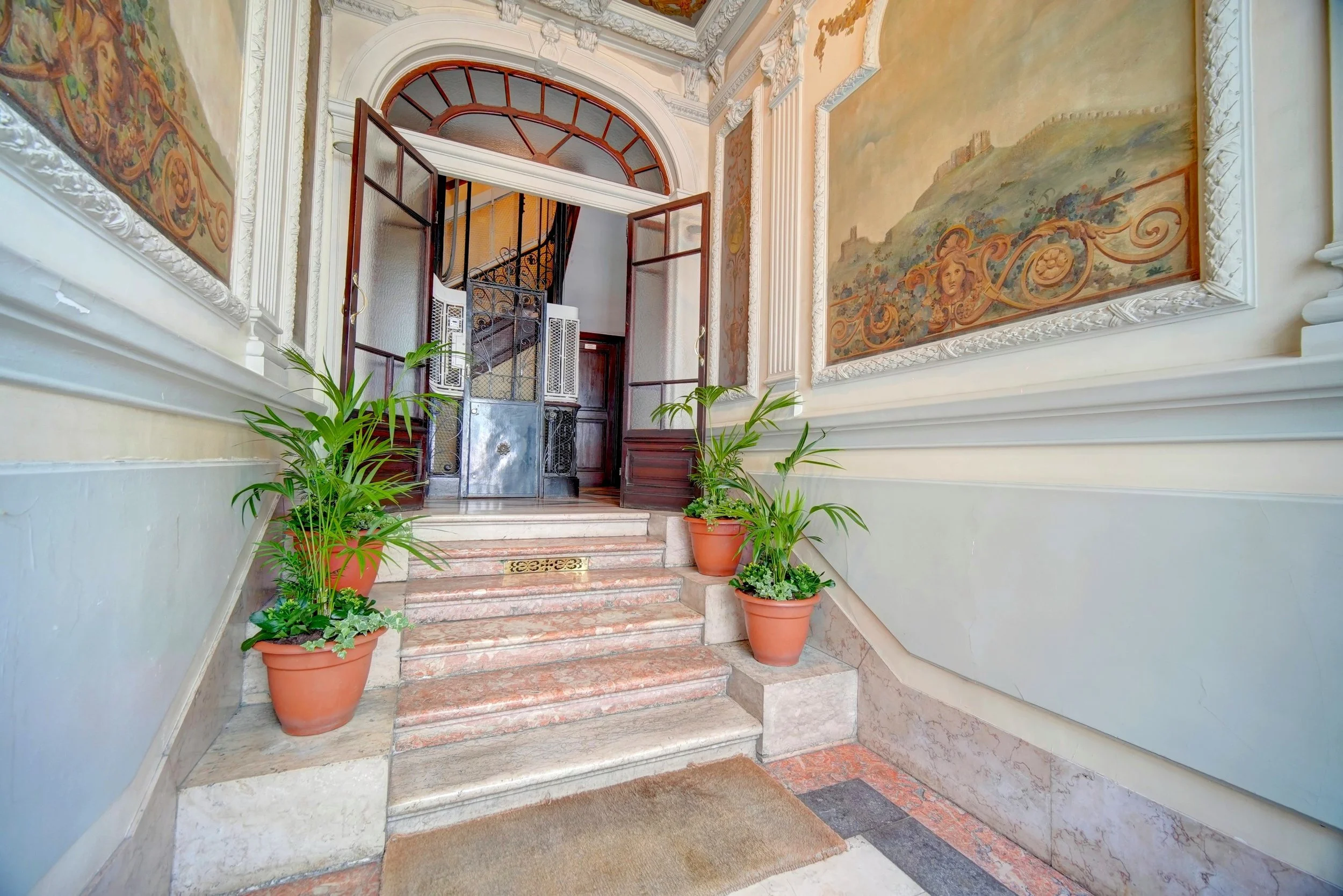 Interior view of a building entrance with marble stairs, potted plants on either side, decorative wall paintings, and an open door leading to a staircase.