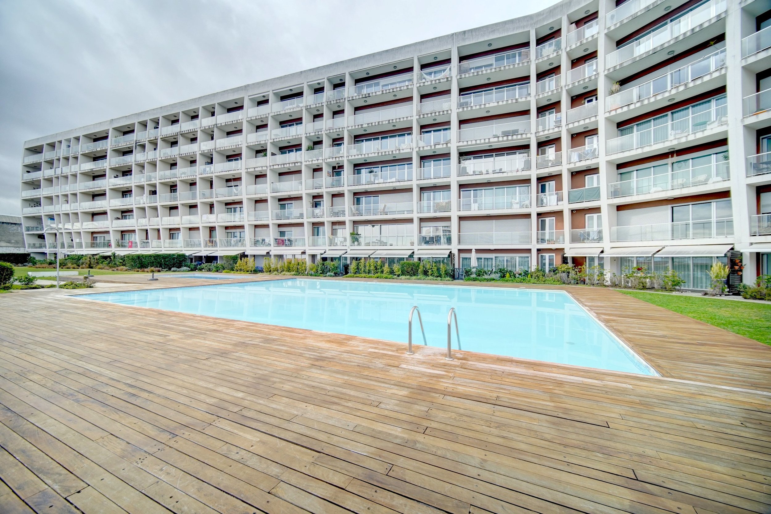 Exterior view of a modern apartment complex with a swimming pool in the foreground, surrounded by wooden decking and greenery.