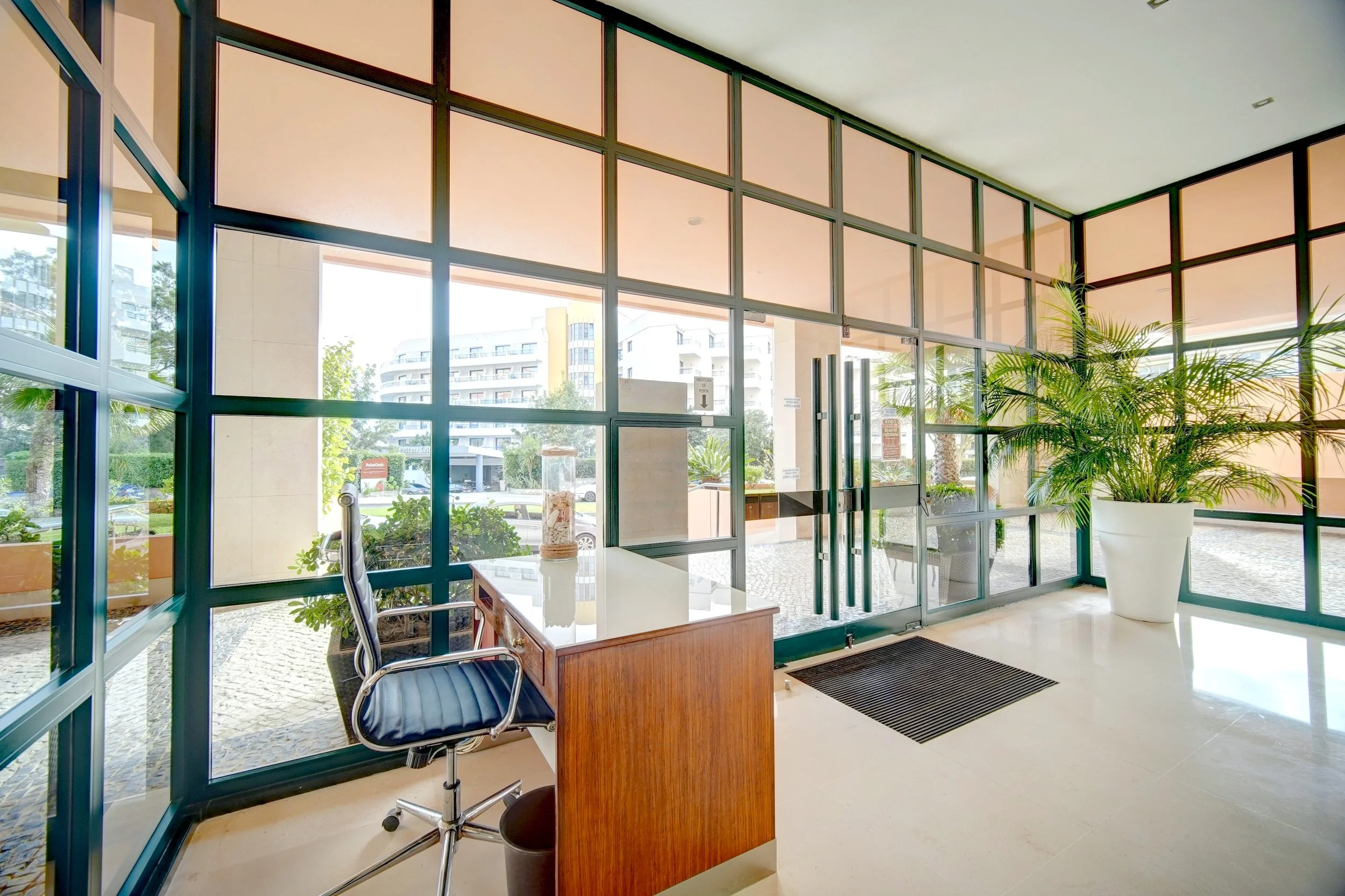Modern building lobby with large glass windows, a wooden desk with a chair, a tall potted plant, and outside visible urban scenery