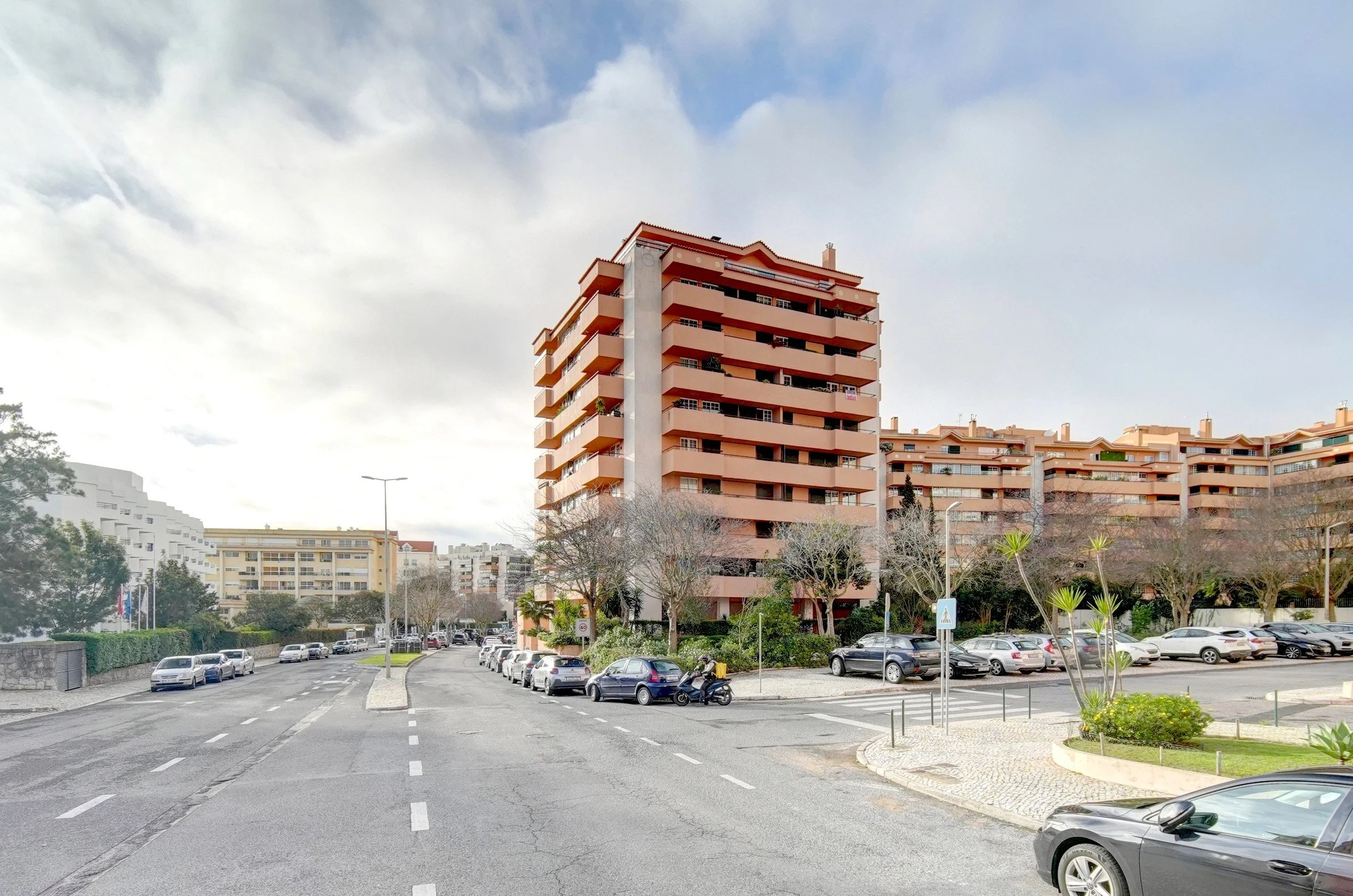 Urban street scene with multi-story orange and beige residential buildings, parked cars, trees, and cloudy sky.