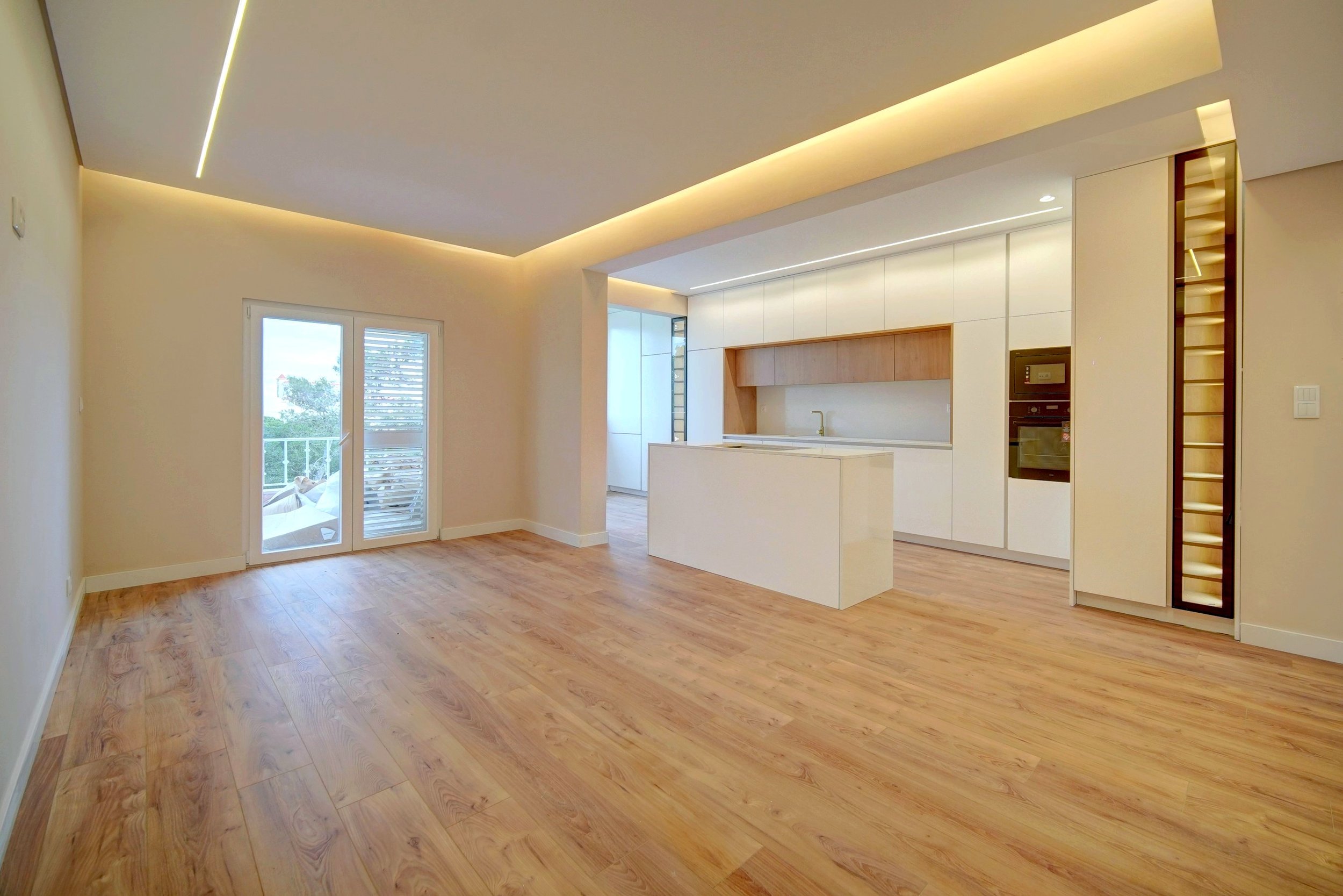 Empty modern kitchen and living area with light wood flooring, white walls, and white cabinetry with wooden accents. Includes a sliding glass door leading to a balcony.