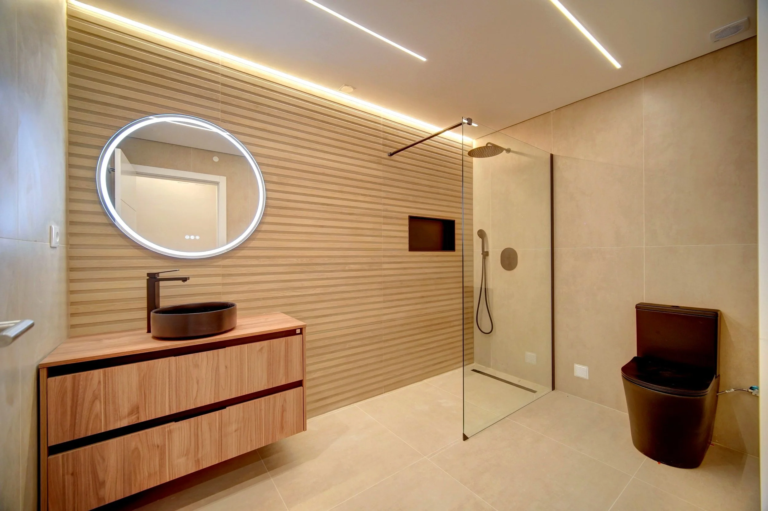 Modern bathroom with a wooden vanity, round black sink, illuminated round mirror, walk-in shower with glass partition, beige tiled walls and floors, and a sleek black toilet.