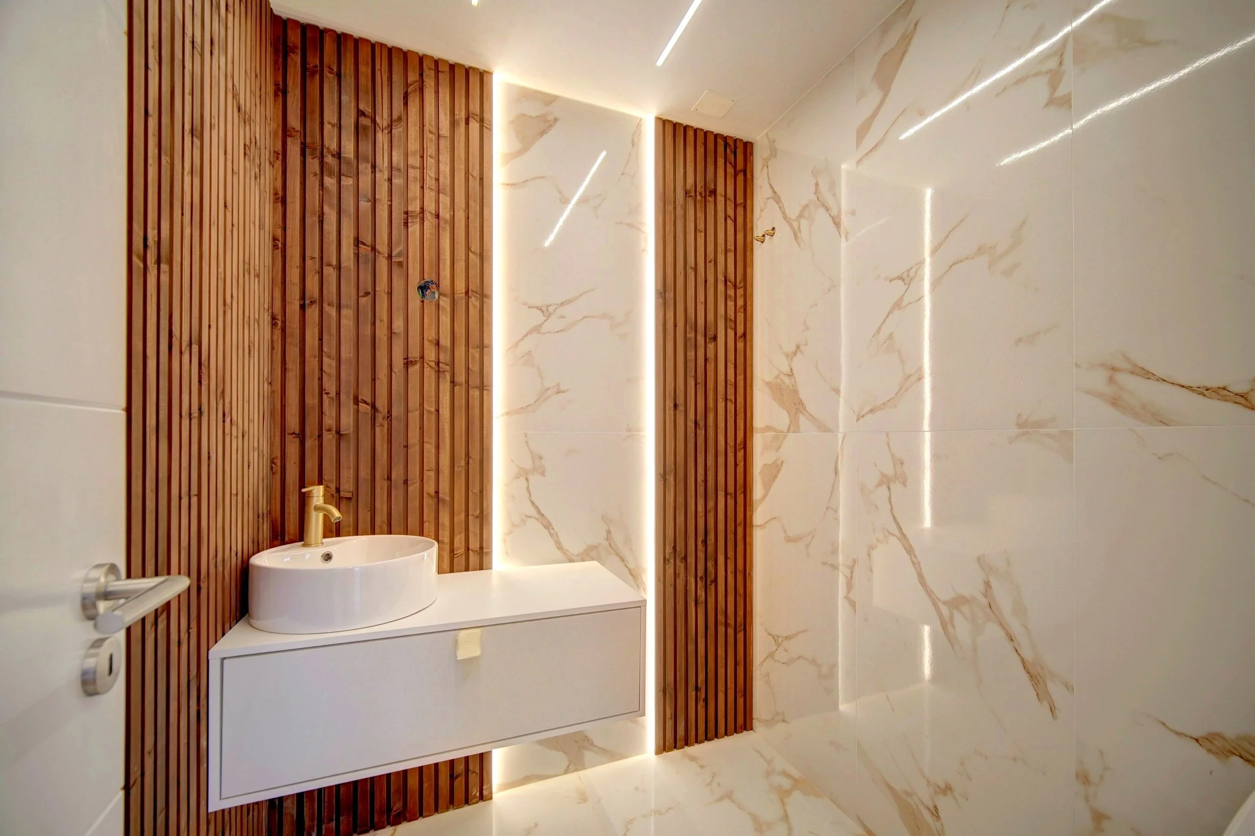 Modern bathroom with marble walls and wood paneling, featuring a white sink with gold faucet above a white cabinet, and LED strip lighting along the walls.