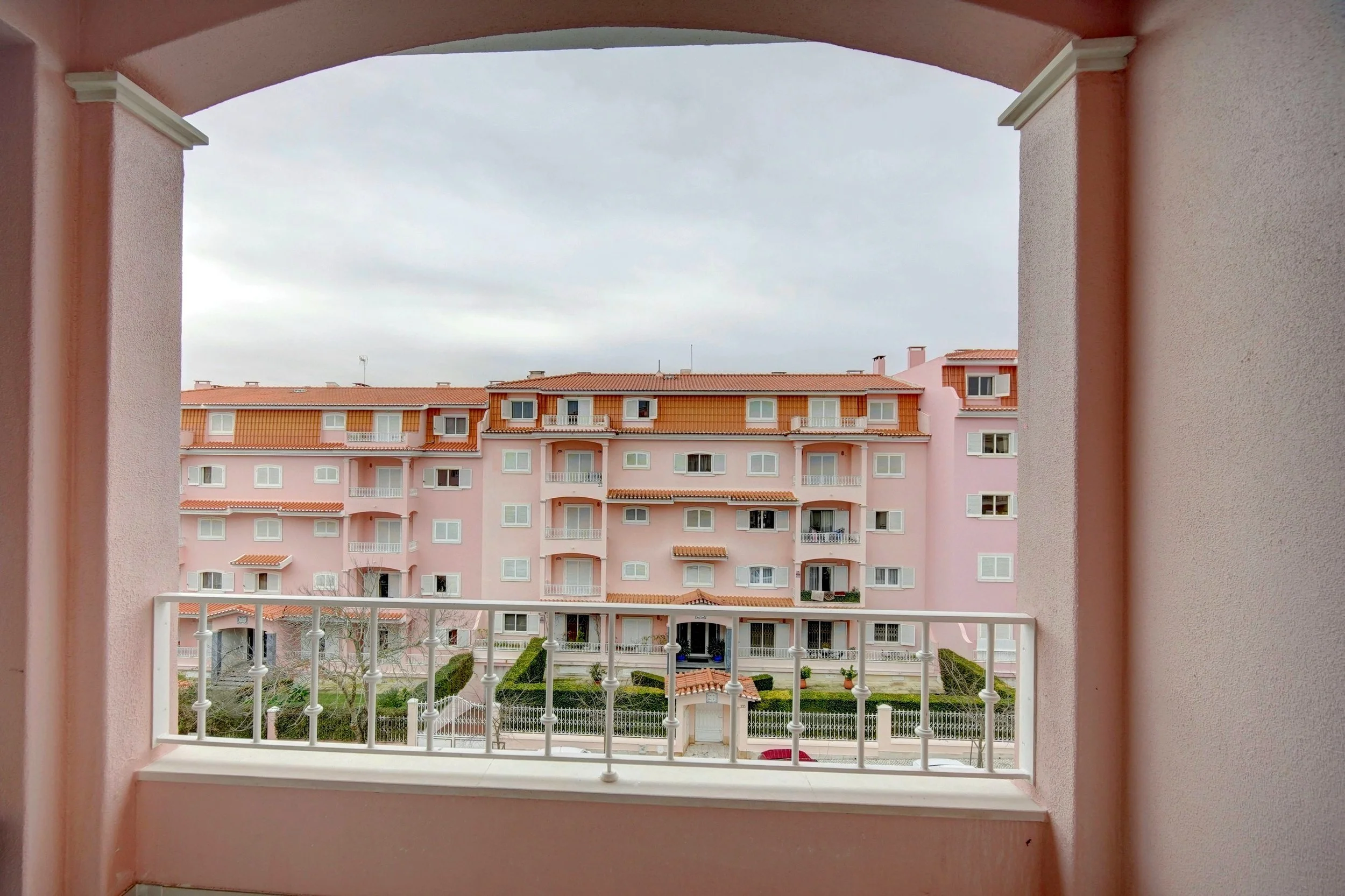 View of a pink residential building with multiple balconies and windows, seen through a balcony opening with a white railing, under an overcast sky.