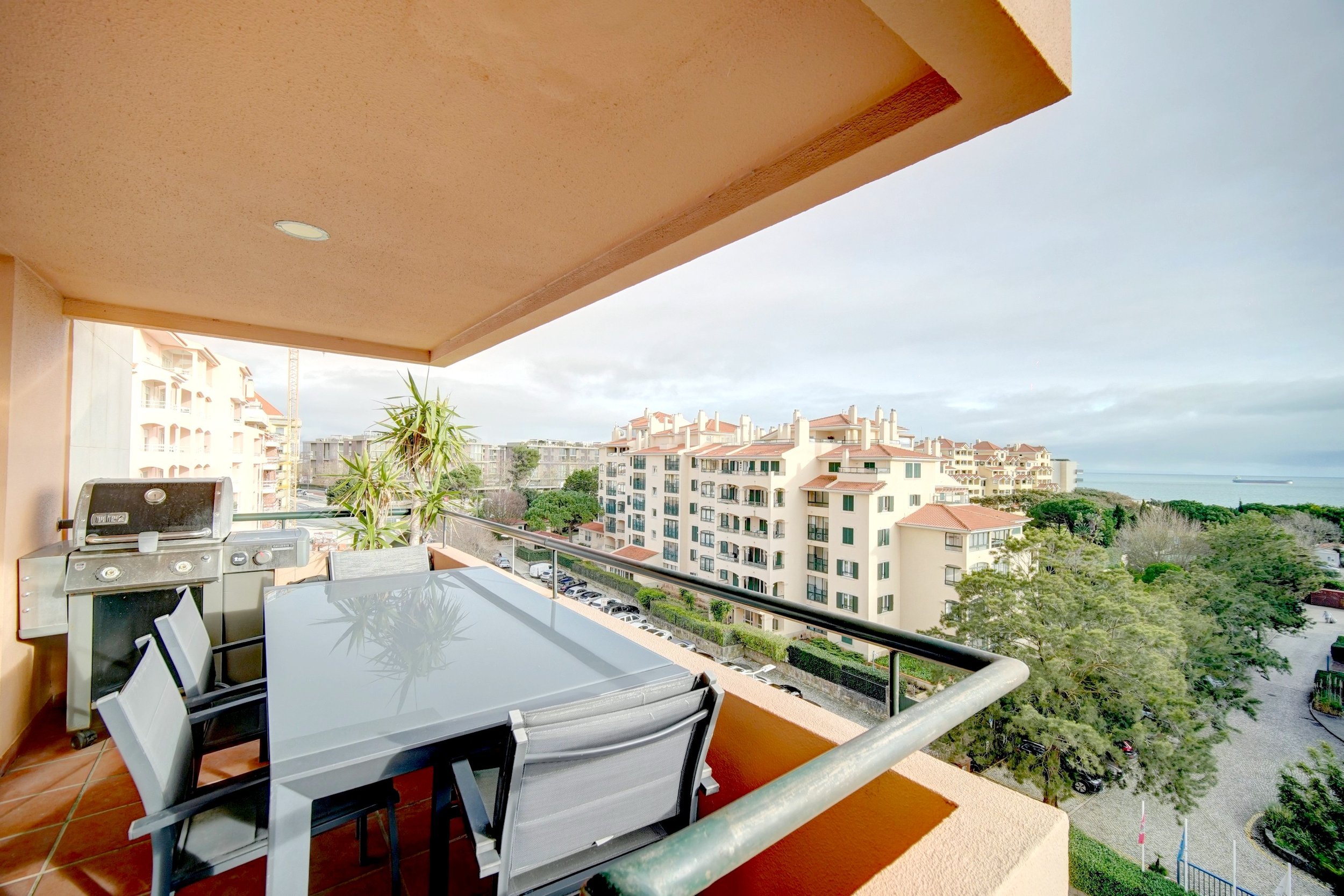 Balcony with a glass-top table, chairs, a barbecue grill, and potted plants overlooking residential buildings, trees, and a distant view of the ocean with a cargo ship.