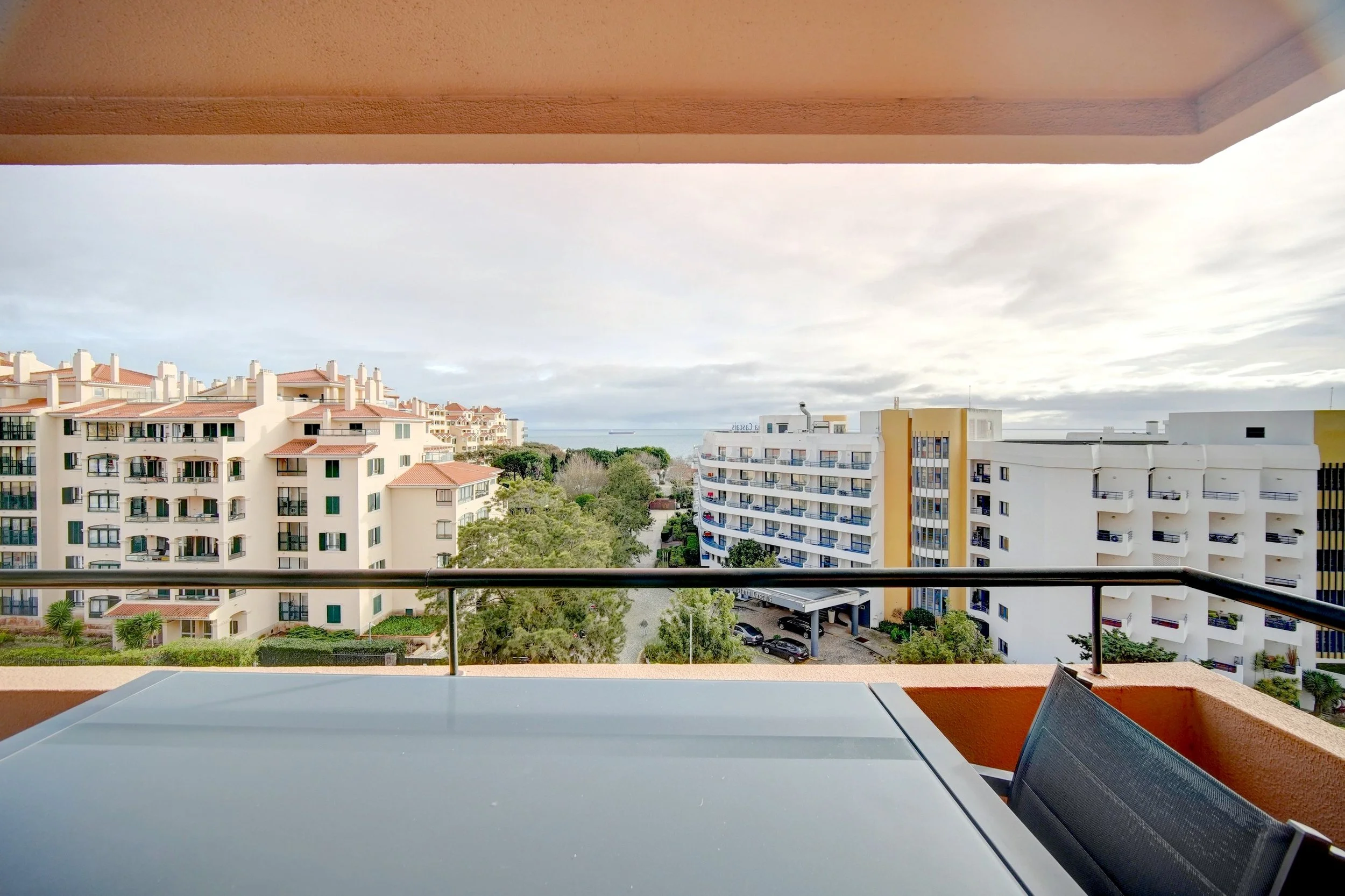 Balcony view of apartment buildings, trees, and a cloudy sky, with a table and chair in the foreground.