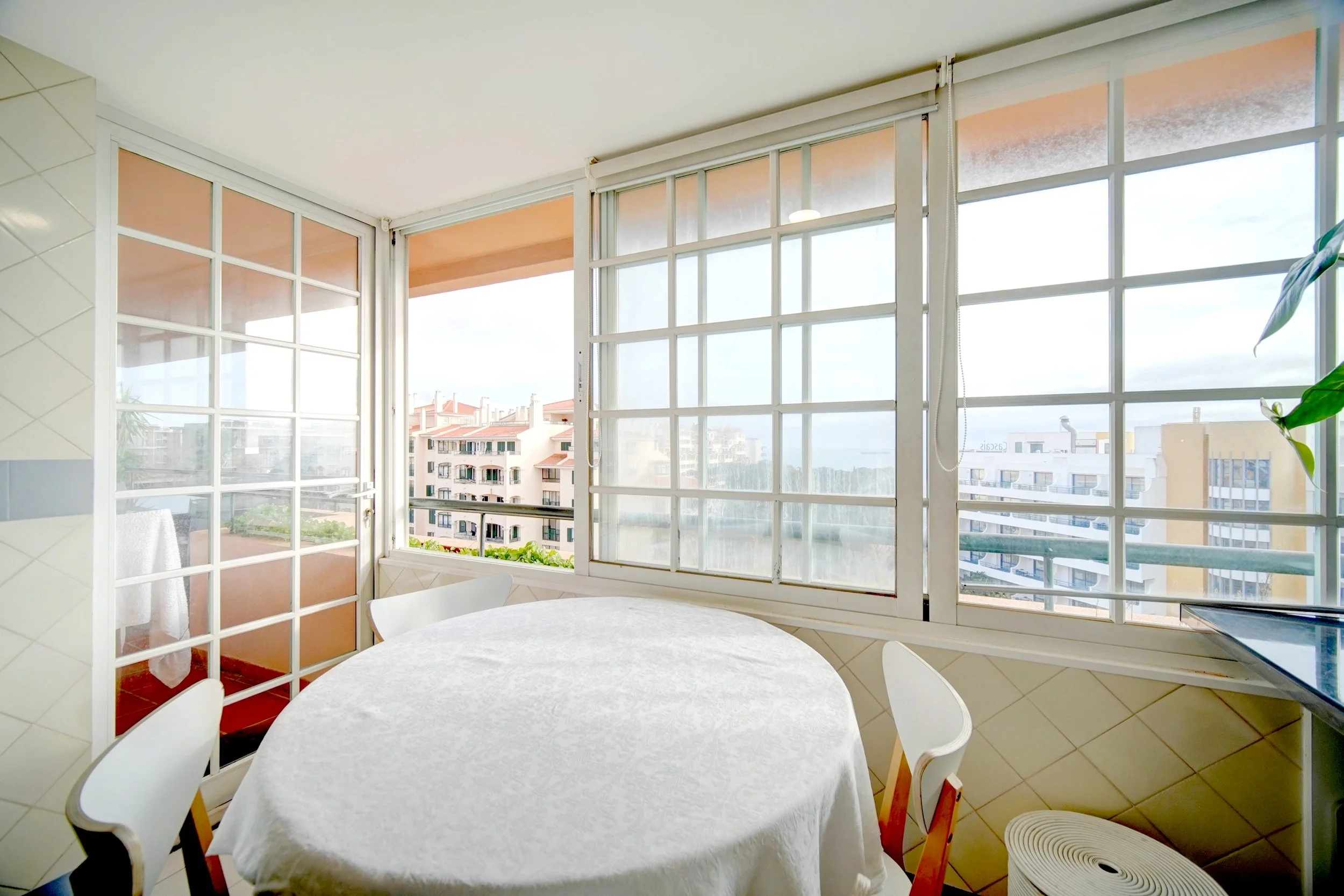 Bright dining area with a round table covered in a white tablecloth, surrounded by white chairs, enclosed by large windows showing an urban skyline with multiple apartment buildings.