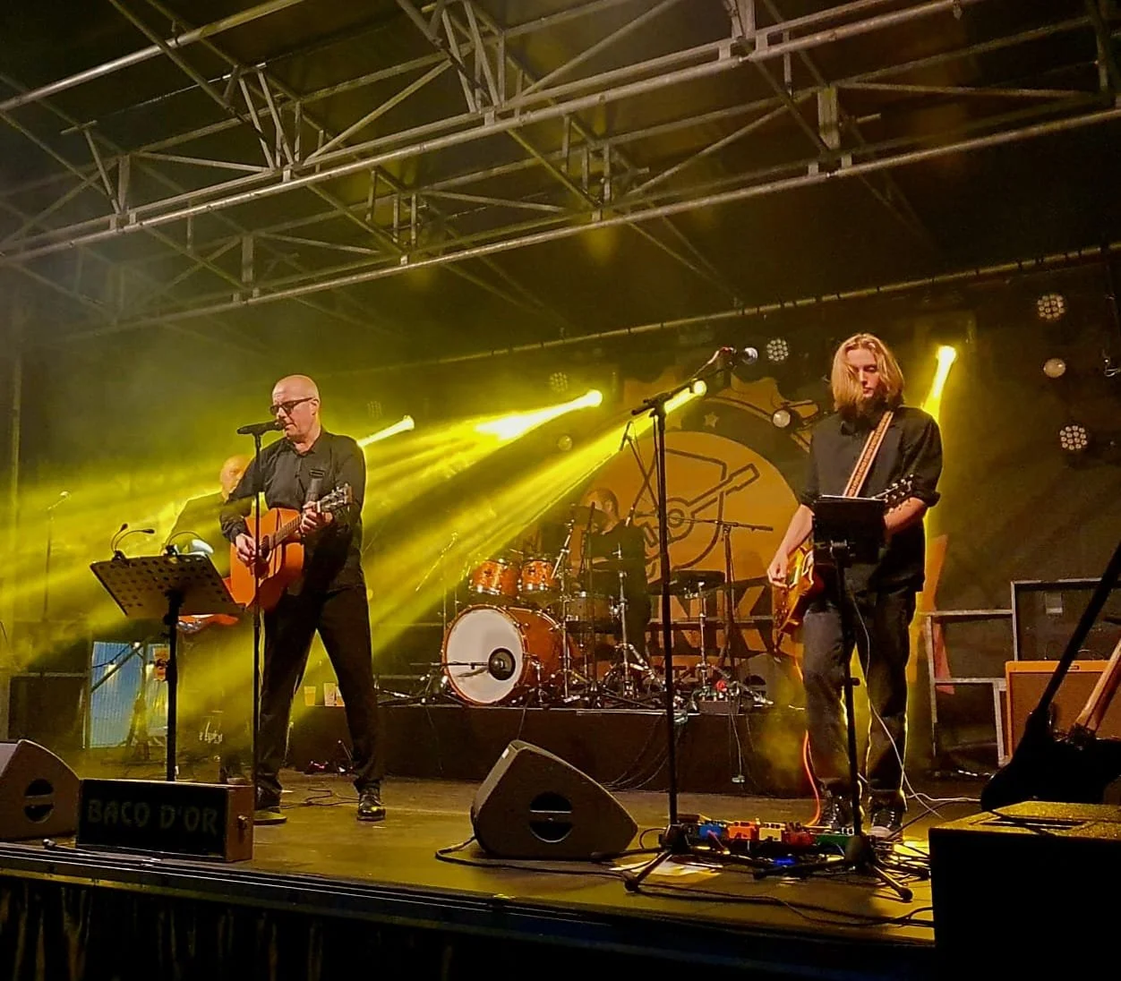 Een muzikale band performing op een podium met gitaar en drums, verlicht door gele podiumlichten.