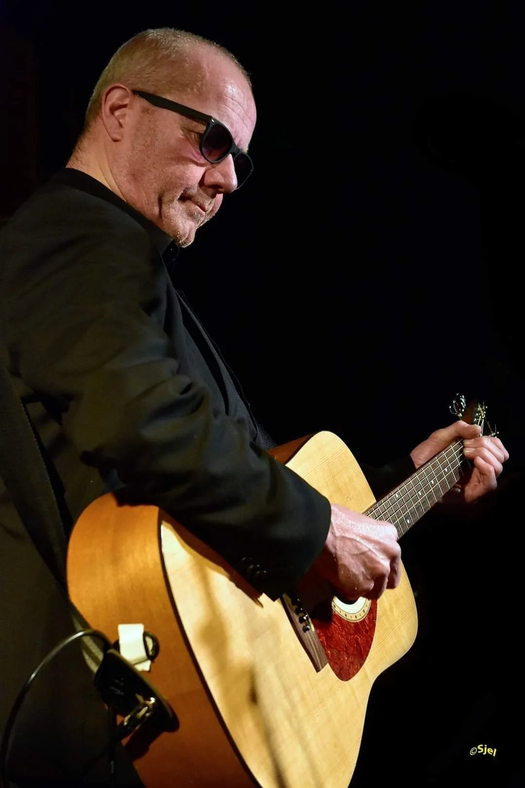 Man met zwart jasje en brillen speelt gitaar op een donkere achtergrond.