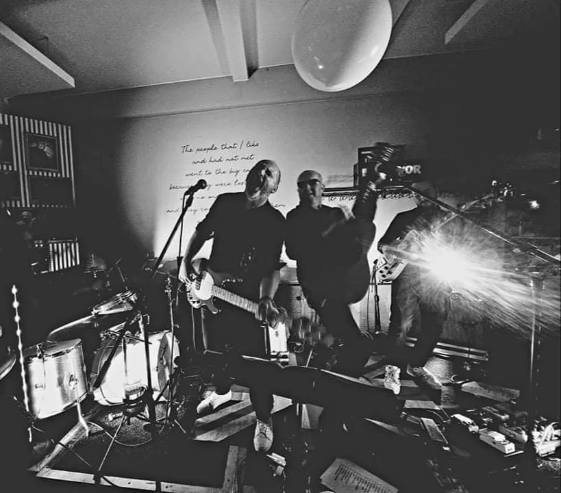 Een band speelt live muziek in een donkere ruimte, met drie muzikanten die gitaar en microfoon gebruiken, een drumstel en andere muziekinstrumenten op de achtergrond, met een lichtstralend effect op de rechterkant.